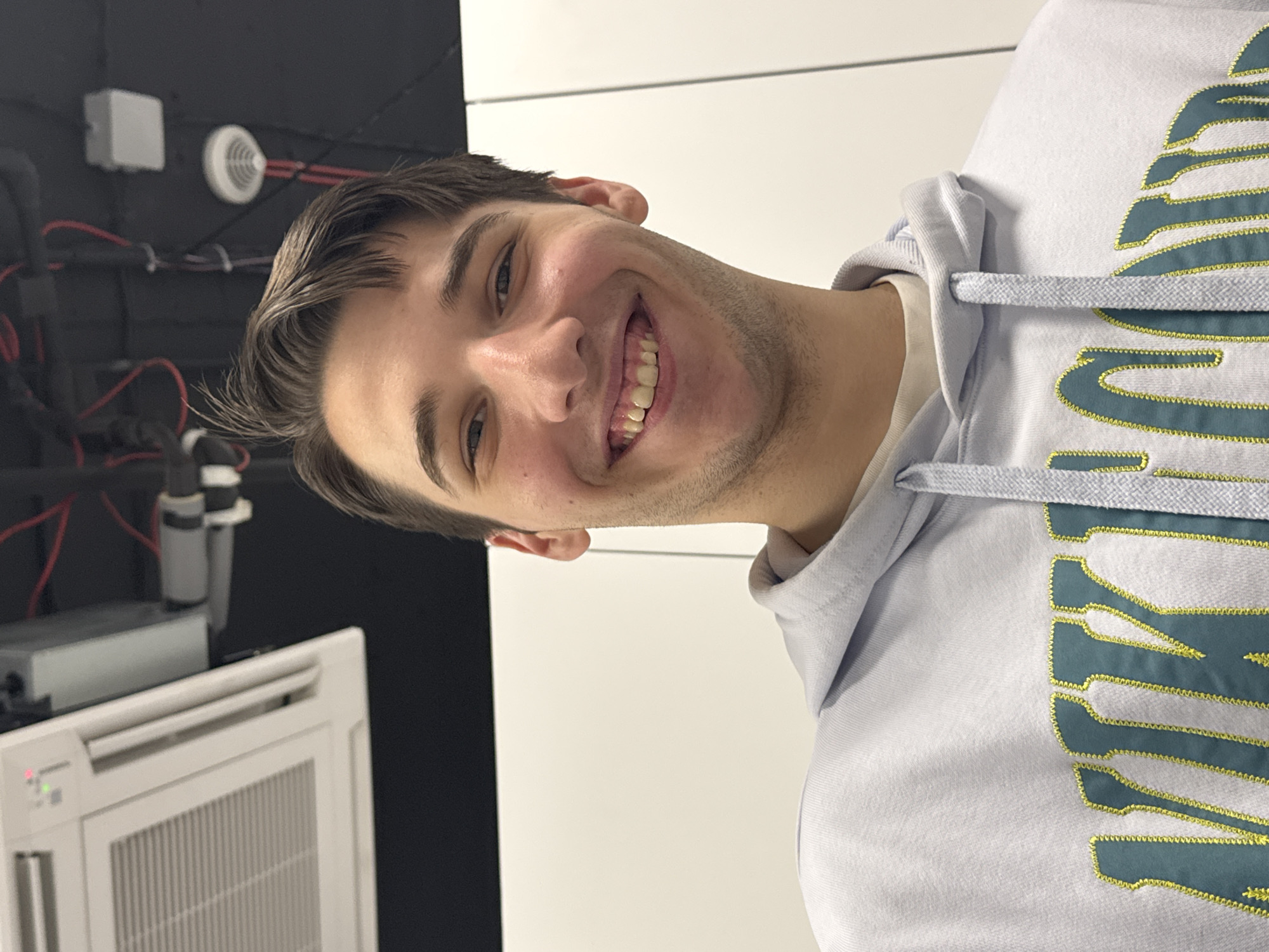 A young man is smiling towards the camera, wearing a light blue hoodie. He has short brown hair and a light complexion. Behind him is a black wall with pipes, a white box, and a smoke detector. There is also a white air conditioning unit. To his right is a white wall. The location is not provided in the image.