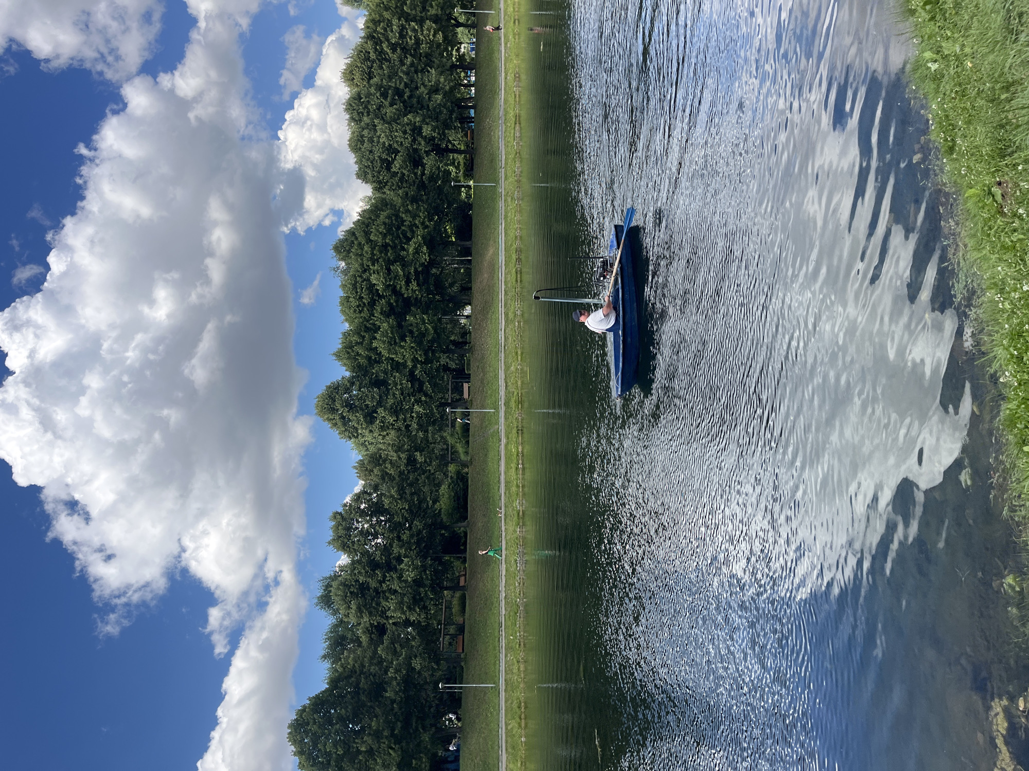 In a serene outdoor scene, a person is seated in a small blue boat on a calm body of water, holding a paddle. The boat is positioned in the middle of the water, with a reflection of the sky and clouds visible on the surface. To the left of the water, a lush green grassy area stretches out, lined with a row of tall, dense trees. Beyond the trees, a clear blue sky is dotted with fluffy white clouds. A pathway runs along the edge of the grass, and a few indistinct figures can be seen further down the path. The lighting suggests it is daytime, likely a clear and pleasant day. The overall impression is one of tranquility and outdoor recreation in a park-like setting, possibly in Moscow, Russia given the context.