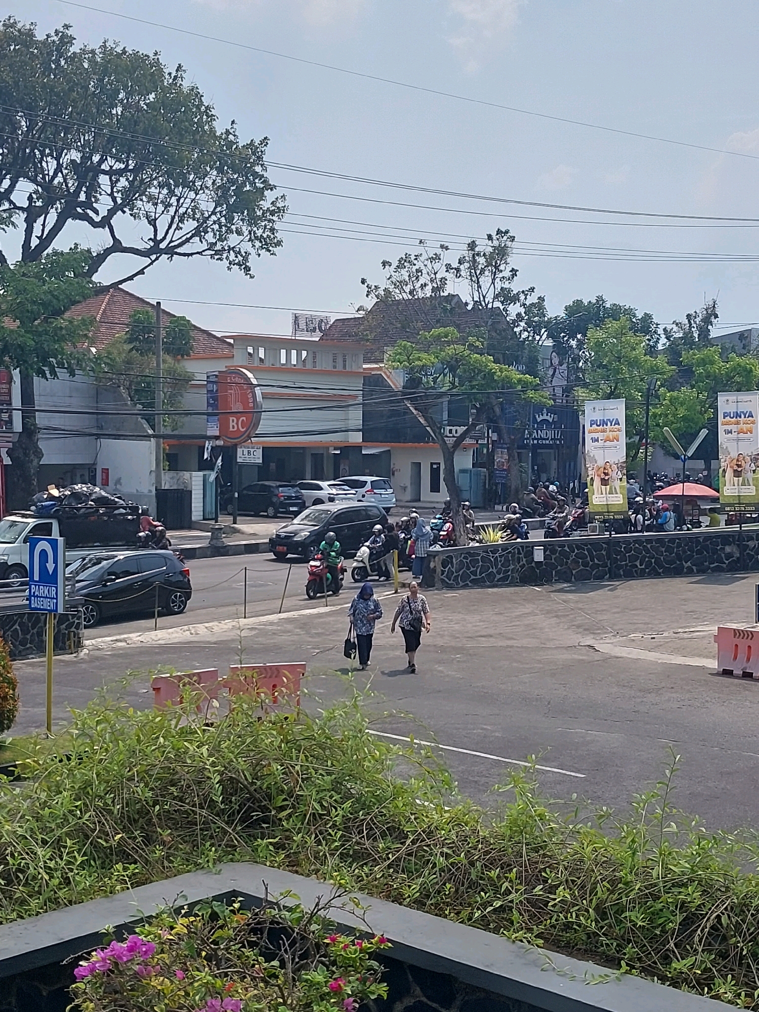 The image depicts a street scene in Malang, Indonesia, likely in a commercial area. In the foreground, a lush green bush with some pink flowers is visible. Beyond the bush, a paved road curves, with several vehicles, including cars, motorcycles, and a truck filled with garbage, present. Two women are walking on the sidewalk, while several people are gathered around parked motorcycles.
Buildings line the street, with signage indicating businesses. One sign displays "LBC" in a circular logo, and another building has "LEG" and "ANDITU" visible. Large banners, possibly advertising, are also displayed, with text that includes "PUNYA BISNIS KOS". Overhead power lines crisscross the sky, which is clear and bright, suggesting a sunny day. The architecture of the buildings appears to be a mix of modern and traditional styles, with tiled roofs visible on some structures. The overall impression is a bustling urban environment.