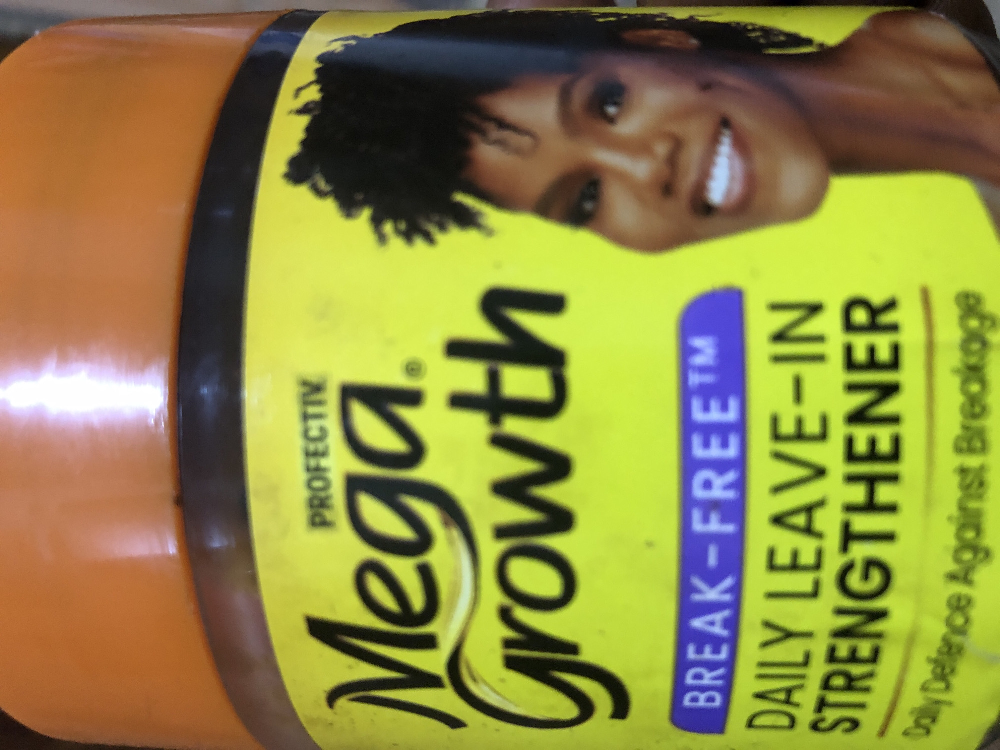 The image shows a close-up of a jar of "Mega Growth" hair product, likely a leave-in strengthener. The jar has an orange lid and a bright yellow label. The label features the brand name "Mega Growth" in large, black, stylized letters. Above "Mega Growth" is the word "PROFECTIV" in smaller, black text. To the right of the text, a portion of a Black woman's face is visible; she is smiling and has short, curly hair. Below "Mega Growth", the text "BREAK-FREE" appears in a purple bar, followed by "DAILY LEAVE-IN STRENGTHENER" in large, black text. At the bottom of the label, smaller text reads "Daily Defence Against Breakage". The background is blurred and appears to be indoors. There is no visible indication of the city of Kabong, Nigeria, within the image itself.