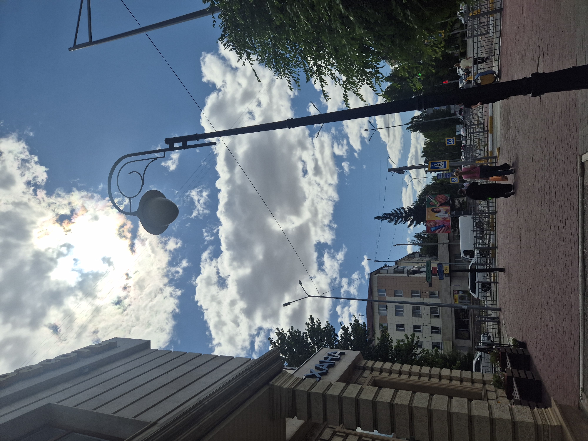 The image captures a street scene in Kokshetau, Kazakhstan, under a partly cloudy sky with the sun partially obscured by clouds. In the foreground on the left, a building with a stone facade and ornate detailing is visible. A sidewalk made of red brick pavers runs in front of the building, featuring benches and planters. A prominent black lamppost with an antique-style lamp stands in the middle of the sidewalk.
Further down the street, there are multi-story buildings, trees, and road infrastructure, including streetlights, traffic lights, and pedestrian crossings. Several vehicles, including cars and a white van, are present on the road. A few pedestrians are walking on the sidewalk. The overall impression is that of a typical urban environment on a bright day.