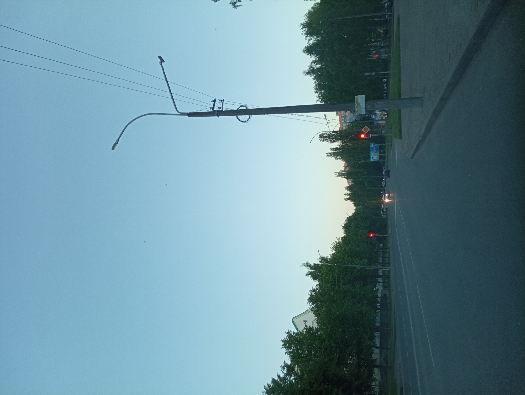 The image depicts a view of a street in Mahilyow, Belarus, likely taken at dusk or dawn due to the soft lighting and clear, pale blue sky. A wide, paved road with white lane markings stretches into the distance, bordered by a sidewalk and a row of mature, green trees on the left. On the right side of the road, a concrete lamppost with multiple overhead wires stands prominently, supporting two streetlights. A red traffic signal is visible ahead on the left, and another on the right. Several cars are present on the road, some with their headlights illuminated. Buildings, including what appears to be a white structure with a green roof on the far left, are partially visible behind the trees. The overall scene suggests a quiet urban environment.