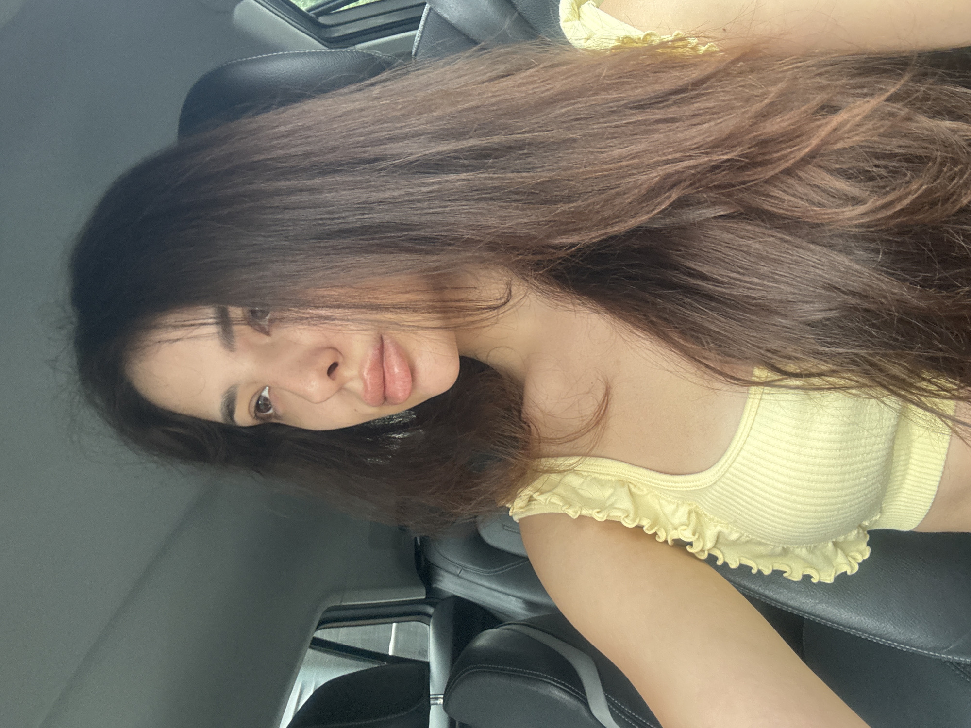 A young woman with long, dark brown hair is captured in a close-up, angled shot, likely a selfie, inside a car. She is positioned reclining in a passenger seat, looking towards the camera with a neutral to slightly pouting expression. Her hair cascades over her shoulder and arm, appearing thick and slightly wavy. She is wearing a light yellow, ribbed crop top featuring delicate ruffles along the hemline and sleeve edges.
The setting is the interior of a modern vehicle, characterized by dark, possibly leather, car seats and headrests. Bright daylight streams in from a visible sunroof or window above, indicating it is daytime. A seatbelt is partially visible across the lower right of the frame. The overall scene is well-lit, highlighting her features and attire against the car's interior. No visible text is present. This scene takes place in Plovdiv, Bulgaria.