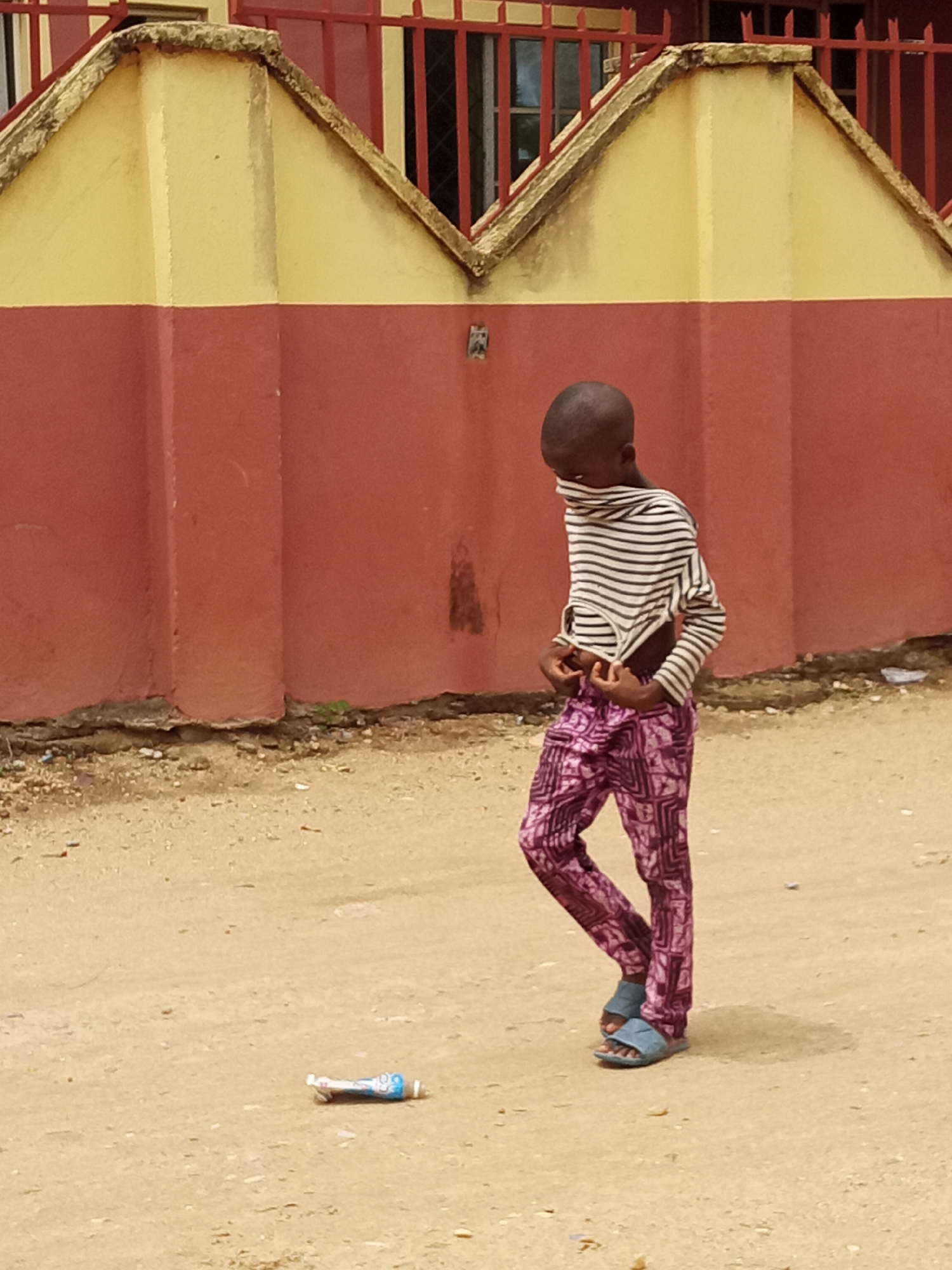 A child stands in a sandy area in the city of Unknown, Unknown. The child, who has a shaved head, wears a long-sleeved striped shirt, patterned purple pants, and blue sandals. The shirt is pulled up over the child's mouth. A discarded blue and white bottle lies on the ground near the child's feet. Behind the child is a building with a red and yellow exterior wall and a red metal fence above. The ground is dry and dusty.