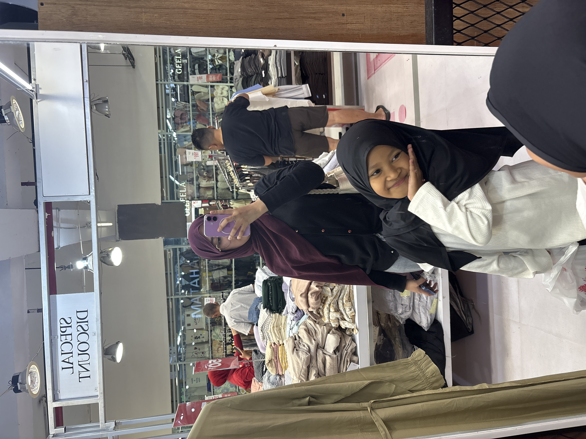 This image, taken in Jember, Indonesia, is a mirror selfie captured inside a bustling clothing or department store. The primary subjects are a young girl and an adult woman, both seen in the mirror's reflection. The young girl, wearing a black hijab and a white long-sleeved top, is positioned prominently in the reflection's foreground. She smiles broadly with both hands playfully touching her cheeks, conveying a cheerful and happy emotion. Behind her, the adult woman, dressed in a maroon hijab and a dark top, holds a purple smartphone to take the picture, with part of her face visible.
The background, also reflected in the mirror, shows a retail environment with numerous clothing racks filled with various garments, including shirts, pants, and what appear to be hijabs. A man in a black t-shirt and shorts is visible in the reflection, browsing clothes on a rack, while other customers can be faintly seen in the distance.
Notable details include bright overhead light fixtures illuminating the store. A white sign hanging from the ceiling, visible in the reflection, reads "SPECIAL DISCOUNT" (reversed). Other visible text in the reflection includes "GEELA" on a product display or shelf, and a partial "MATAHARI," strongly suggesting the Indonesian department store Matahari. A price tag displaying "100K" is also discernable. The scene captures a lighthearted moment during a shopping trip, likely daytime or evening, given the indoor setting.
