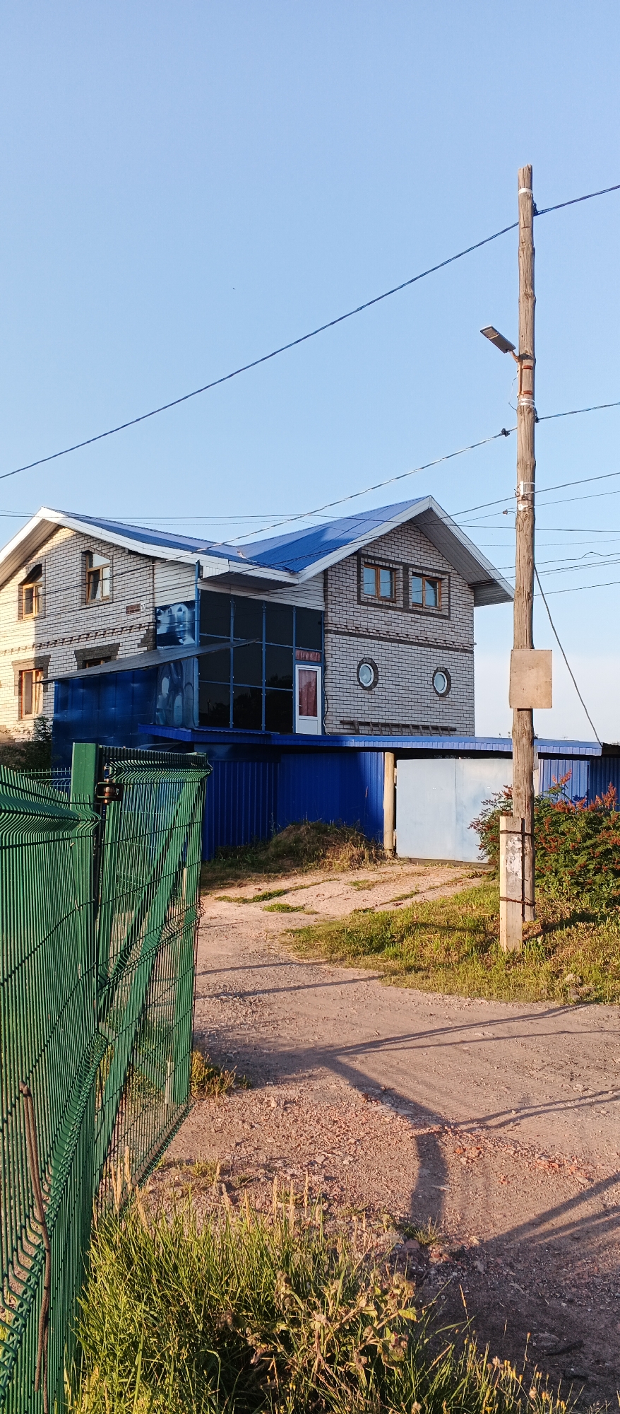 The image depicts a two-story brick building with a blue roof, situated in Izhevsk, Russia. The building features large glass windows on the ground floor, a red door, and circular windows on the upper floor. A green metal fence runs along the foreground of the image, with a dirt road leading to a light blue gate in front of the building. A wooden utility pole with a street lamp stands to the right of the frame, with power lines stretching across the sky. The environment is rural or semi-rural, with overgrown grass and vegetation surrounding the property. The sky is clear and blue, indicating daytime. There are no people or animals visible in the image.