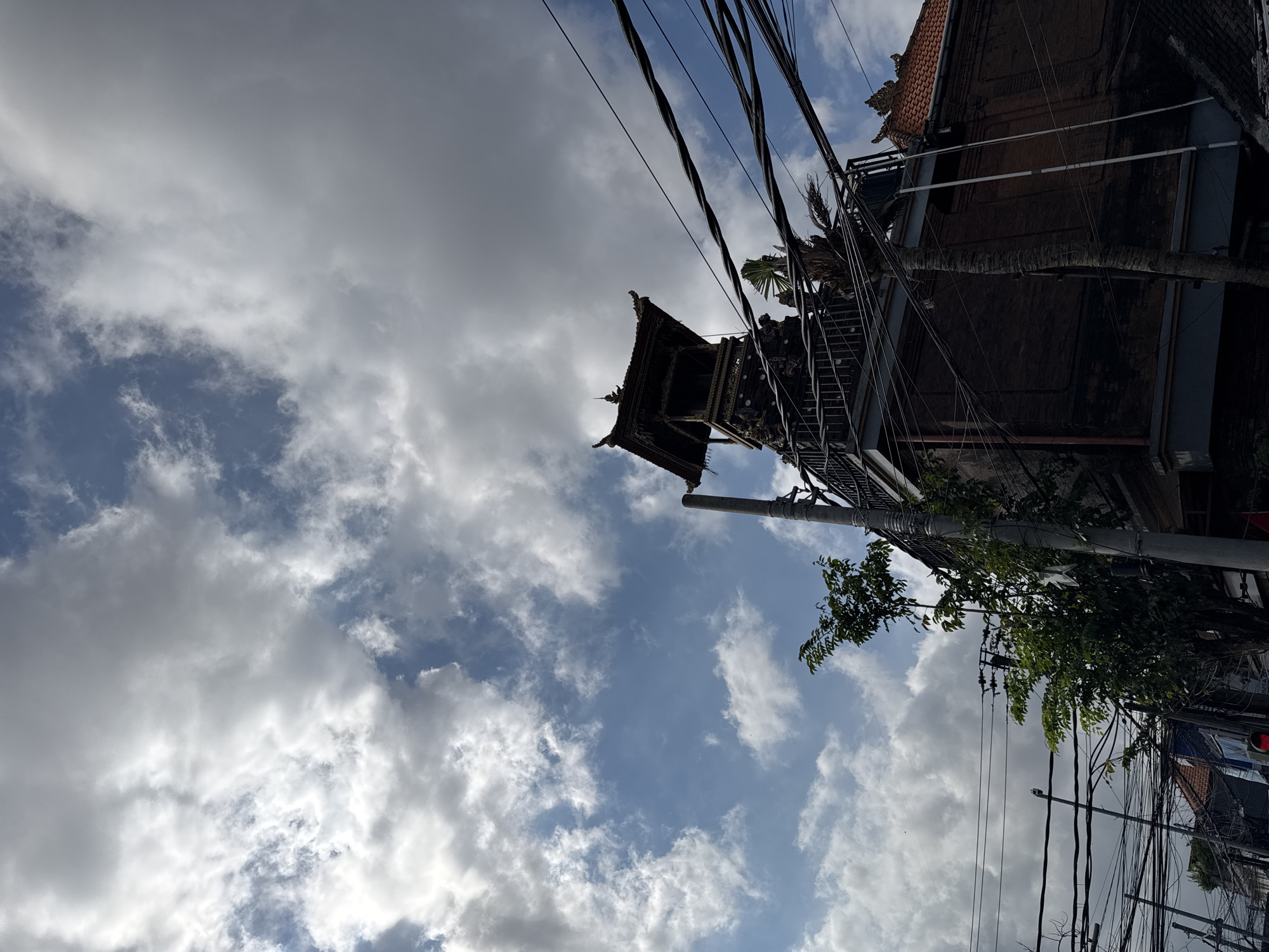 The image captures a view from street level in Denpasar, Indonesia, looking upwards towards the sky and a traditional Balinese structure. A dark, multi-tiered temple-like building with intricate carvings and a roof reminiscent of traditional Balinese architecture is prominently featured on the right side of the frame. Several power lines are strung across the sky, obscuring parts of the view and adding a sense of urban density. Green trees and foliage are visible in the foreground and to the left of the building, contrasting with the man-made structures and the dramatic cloudy sky. The clouds are a mix of bright white and darker grey, suggesting dynamic weather conditions. The architecture of the building suggests it is likely a place of cultural or religious significance within the city.
