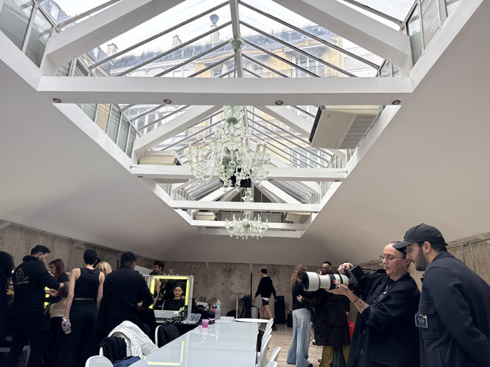 The image depicts an indoor scene beneath a large glass skylight, revealing typical Parisian rooftops and building facades, confirming the location as Paris, France. Within the space, a group of approximately 10-15 individuals is engaged in activities consistent with a photoshoot or event preparation. In the right foreground, a woman with glasses and arm tattoos operates a professional camera with a prominent white telephoto lens, aiming it towards the left. Beside her, a man wearing a black baseball cap and dark jacket with a Stone Island patch observes in the same direction. To the left and mid-ground, several individuals are gathered around long white tables. Two illuminated mirrors are visible on a table, with a woman's reflection seen in one, suggesting hair or makeup application. Other people are standing, walking, and interacting, with one person having their hair styled on the far left. The overall lighting is natural from the skylight. Two multi-tiered chandeliers hang from the skylight structure, and an air conditioning unit is mounted within the framework.