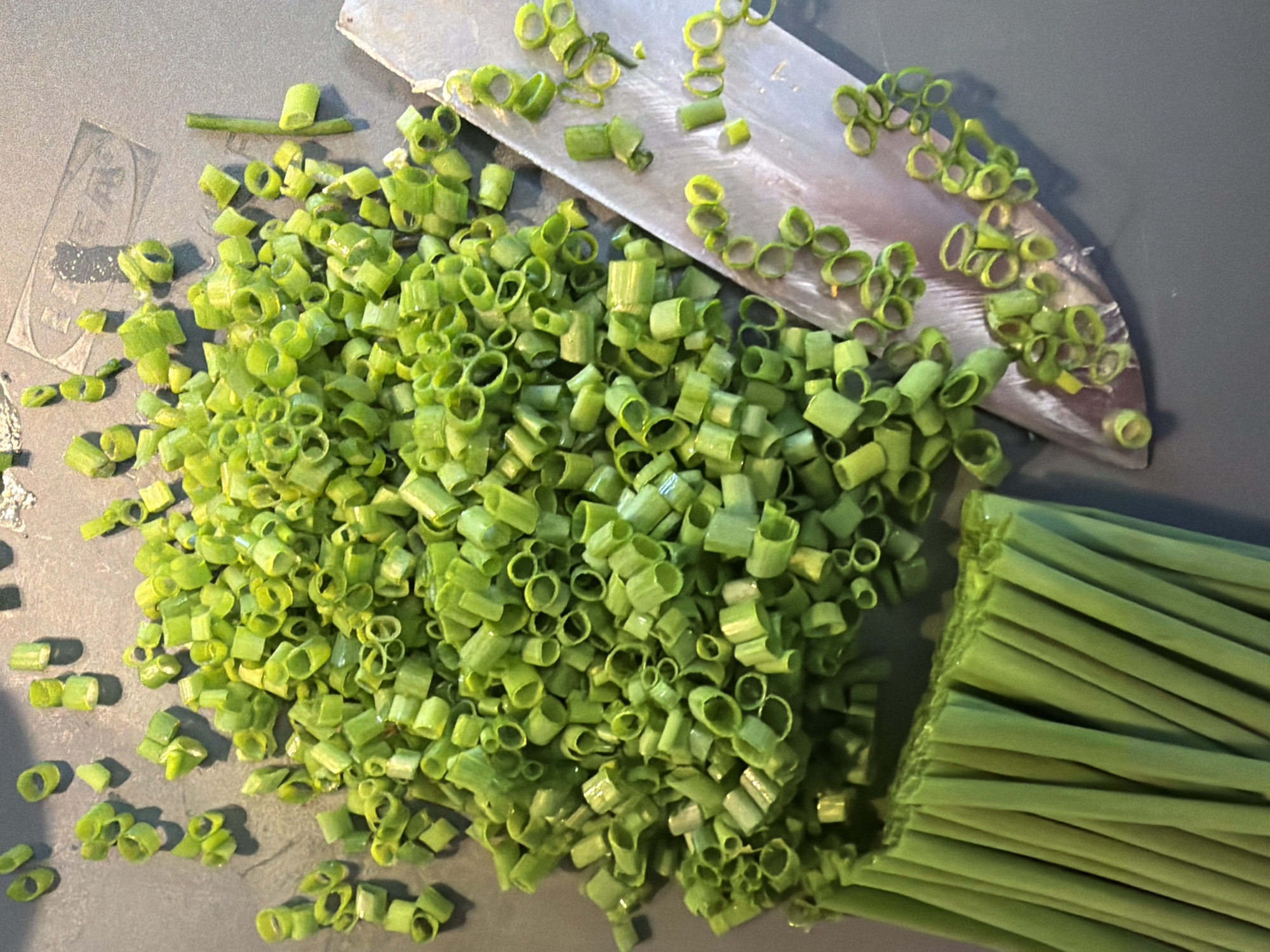 The image depicts a gray cutting board on which chopped green onions are piled, with a knife resting on top of the pile. Unchopped green onions are stacked to the right of the pile. The environment appears to be a kitchen setting. No people are visible. There is no evidence to indicate the location of this image is Saint Petersburg, Russia.