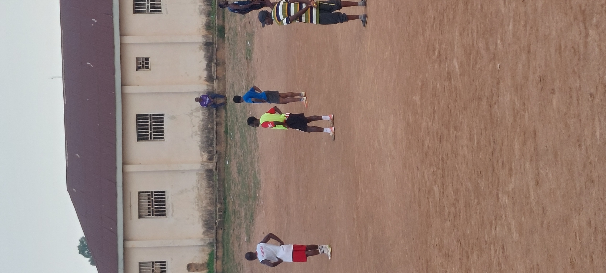 In Bukuru, Nigeria, an outdoor sports field is depicted. A long, light-colored building with a dark, sloped roof stands in the background. The building has multiple barred windows and appears to be aged, with some weathering visible on its exterior. Several individuals are scattered across the dirt field. In the foreground, on the left, a person in a white tank top with "GOMBE" printed on it and red shorts stands with hands on their hips, facing away from the camera. To their right, another person in a red t-shirt and a bright yellow vest with black shorts is looking to the side. Further to the right, a person in a blue t-shirt and dark shorts stands with their hands on their hips, also facing away. In the middle ground, on the right side of the frame, a person wearing a striped t-shirt and dark shorts stands with their back to the camera, holding a yellow object. In the far background, a person in a purple shirt sits on a raised surface in front of the building. The ground is a reddish-brown dirt, with patches of dry grass. The sky is overcast and pale.