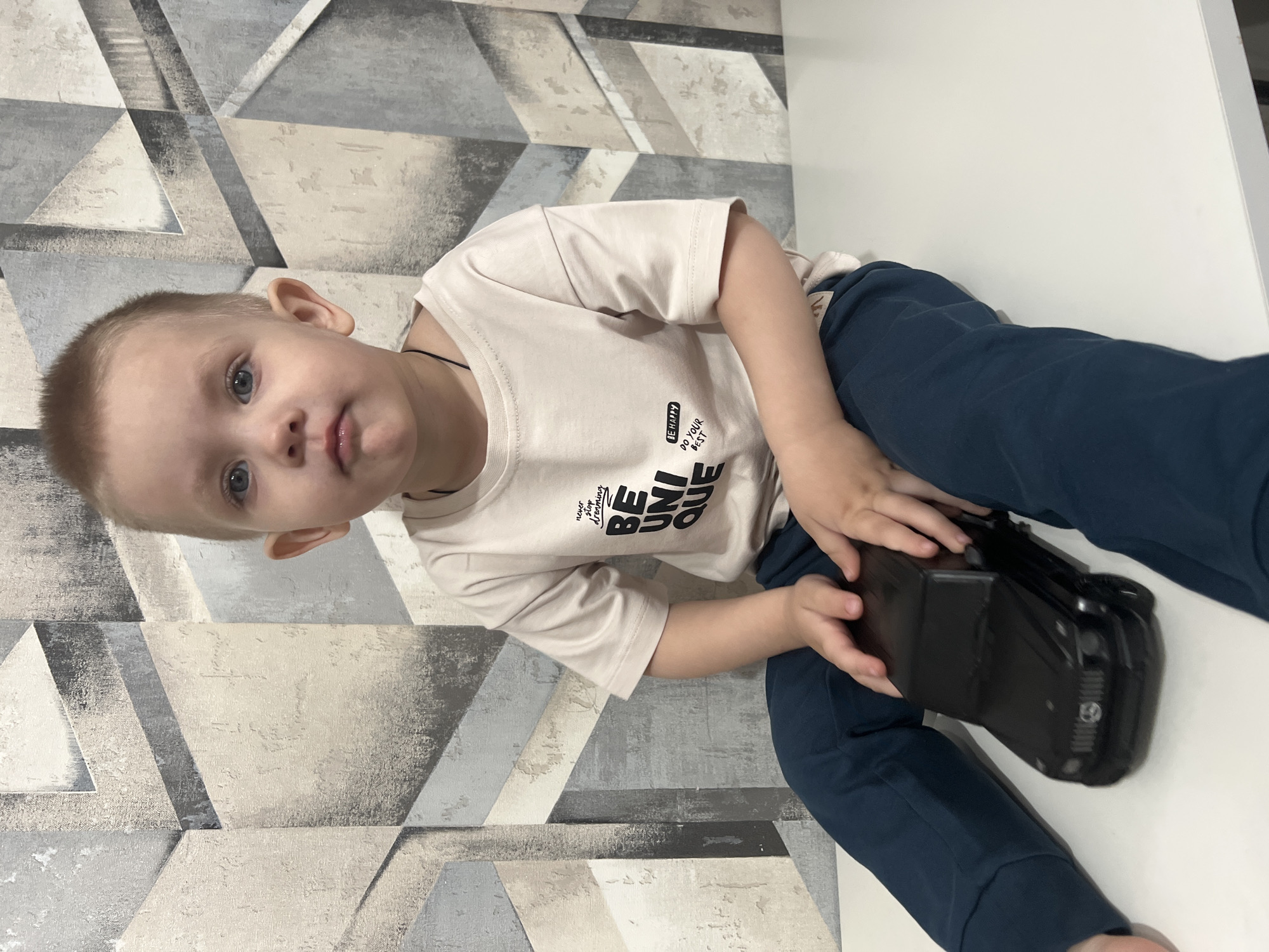 A young boy with short blond hair and blue eyes is sitting on a white surface, holding a black camera. He is wearing a light beige t-shirt with "BE UNIQUE" printed on it and dark blue pants. The background consists of a wall with a geometric pattern of gray and beige. The image is rotated clockwise. The location is Марковский, Russia.