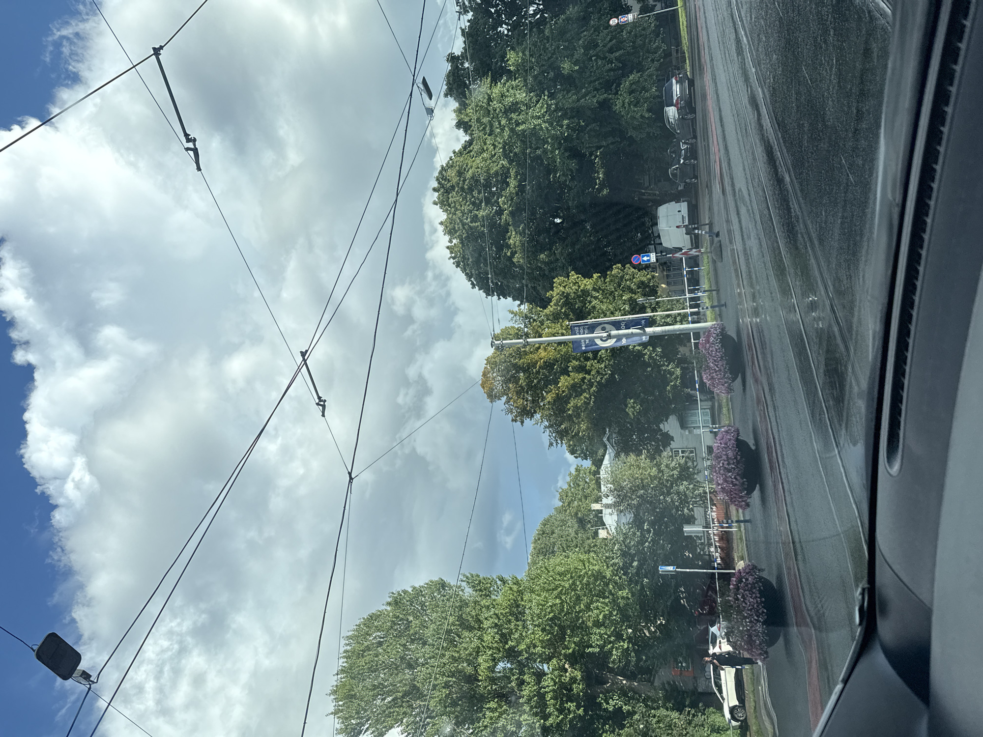 The image captures a view from inside a vehicle in Tallinn, Estonia. The scene is a city street with tram tracks in the foreground, indicating a public transportation route. The road surface appears wet, suggesting recent rain. Large, mature trees line the street, with lush green foliage, some showing hints of autumn color. In the background, buildings are visible behind the trees, and some structures appear to be residential. Overhead, a complex network of tram wires crisscrosses the sky, punctuated by streetlights. Flower planters filled with pink flowers are strategically placed along the roadside, adding a touch of urban landscaping. A few cars are parked or moving on the street, and a person is walking near a white vehicle on the left side of the frame. The sky is partially cloudy with patches of blue sky. The overall impression is of an urban environment with significant greenery and infrastructure for public transport.