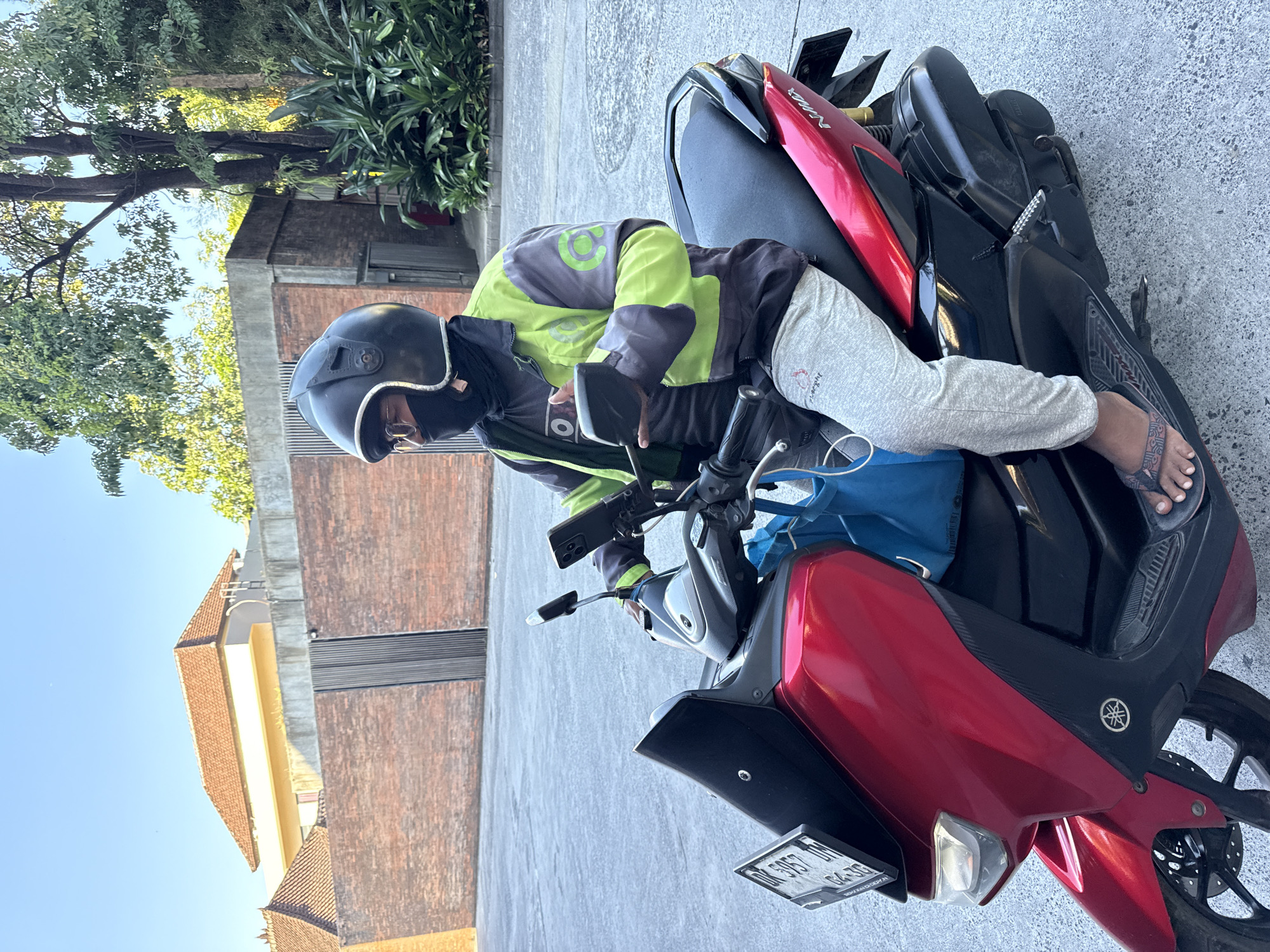 A person wearing a black helmet, black face mask, a dark grey shirt with a lime green and dark grey jacket with green circular logos, and light grey sweatpants sits on a red and black Yamaha NMAX scooter. The scooter has a blue bag hanging from the handlebars and a black phone mount on the right handlebar, holding a black smartphone. The scooter's license plate, which is black with white text, is partially visible. The person's feet are in dark-colored flip-flops. The scooter is parked on a grey asphalt surface. In the background, there is a brick wall with a metal gate, a light-colored building with a red tile roof, and lush green trees and foliage. The scene appears to be outdoors, possibly in a residential or commercial area.