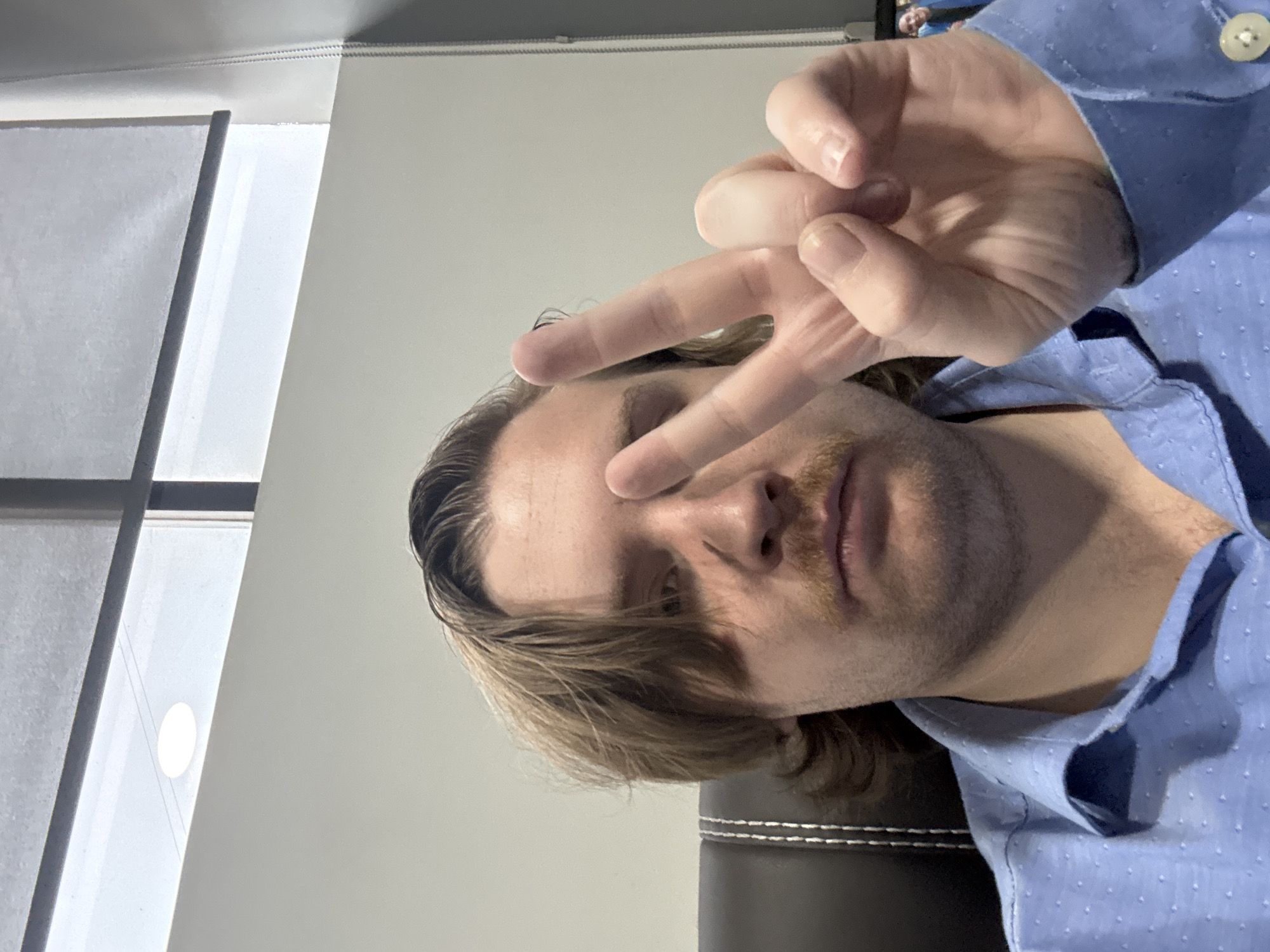 The image shows a close-up, angled portrait of a person indoors in what appears to be Cuautitlán Izcalli, Mexico. The individual, who appears to be male, has light brown hair and a mustache. They are wearing a blue collared shirt with a subtle textured pattern of small white dots and a white button on the cuff of the right sleeve.
The person is posing casually, holding their right hand up in a peace sign (V-sign), with their fingers partially obscuring their face. Their gaze seems directed towards the camera. Part of a dark, possibly leather, chair with white stitching is visible on the lower right side of the frame, suggesting an office or home environment. In the background, on the left, a bright window or vertical blinds are visible, letting in diffused light. The overall lighting is soft, illuminating the subject and the indoor setting. No specific time of day or outdoor weather conditions can be determined from the indoor scene. There is no visible text in the image.