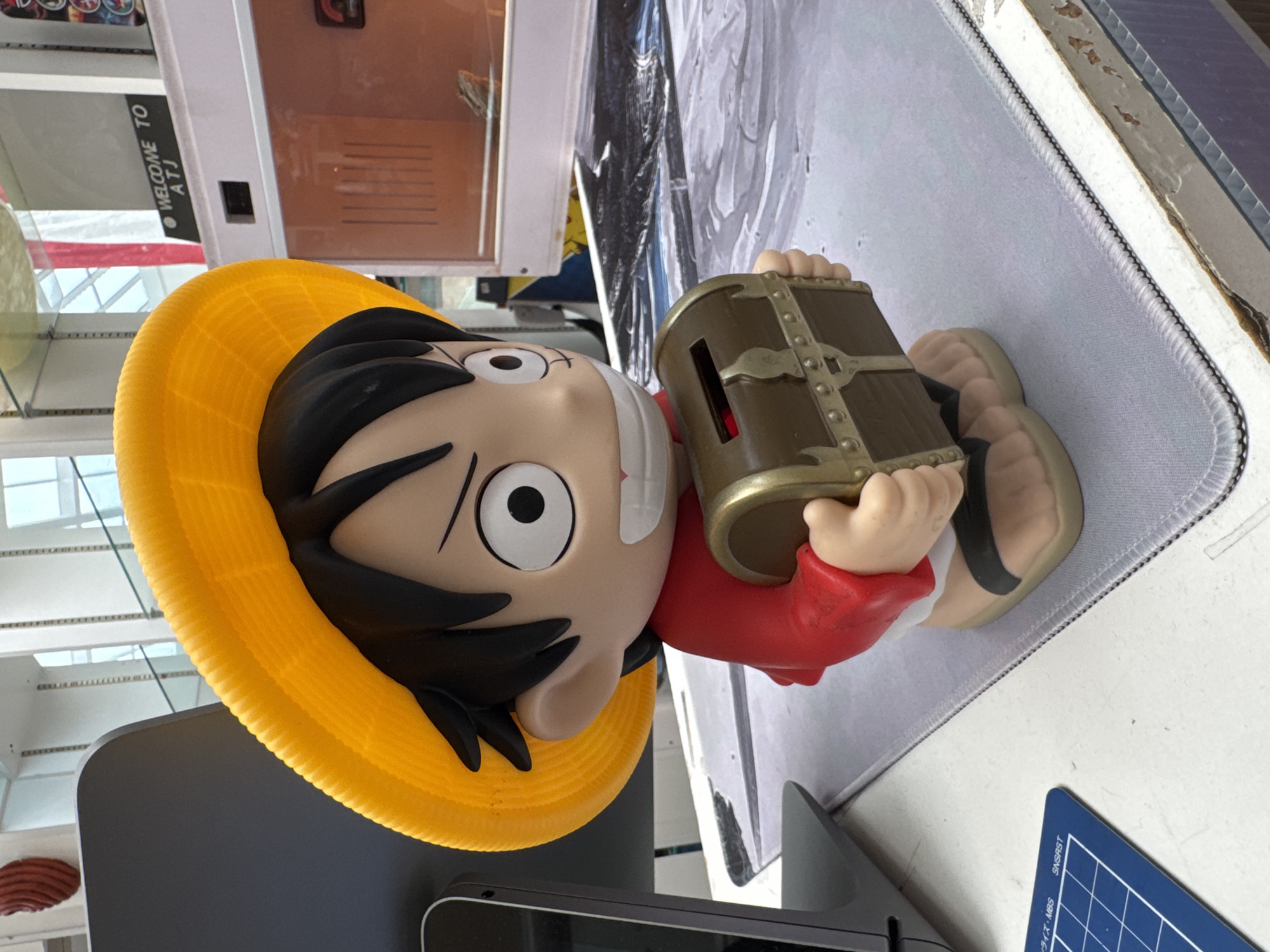 A close-up, eye-level, medium shot captures a cartoon-like piggy bank shaped like the character Monkey D. Luffy from the anime One Piece. The piggy bank is predominantly yellow for the hat, red for the shirt, and beige for the skin and feet, wearing black sandals. It holds a small, brown treasure chest with gold accents, which serves as the coin slot. The character has large, round eyes with black pupils and an open-mouthed smile.
The piggy bank is placed on a light gray mousepad with a textured surface. Behind the character, a large, dark gray, angled object, possibly a monitor or laptop screen, is visible on the left. In the background, a white counter or table supports various items, including a white and beige box with a glass front, inside which a small reptile, likely a lizard or gecko, is perched. A black sign above this box reads "WELCOME TO ATJ". The overall environment suggests an indoor retail or display space, potentially a gift shop or electronics store. The lighting is bright and natural, coming from above, indicating a well-lit interior, possibly with large windows or a skylight. The image composition focuses on the piggy bank as the central subject, with the background providing context for its display. There is no indication of the specific location within El Paso, United States of America.