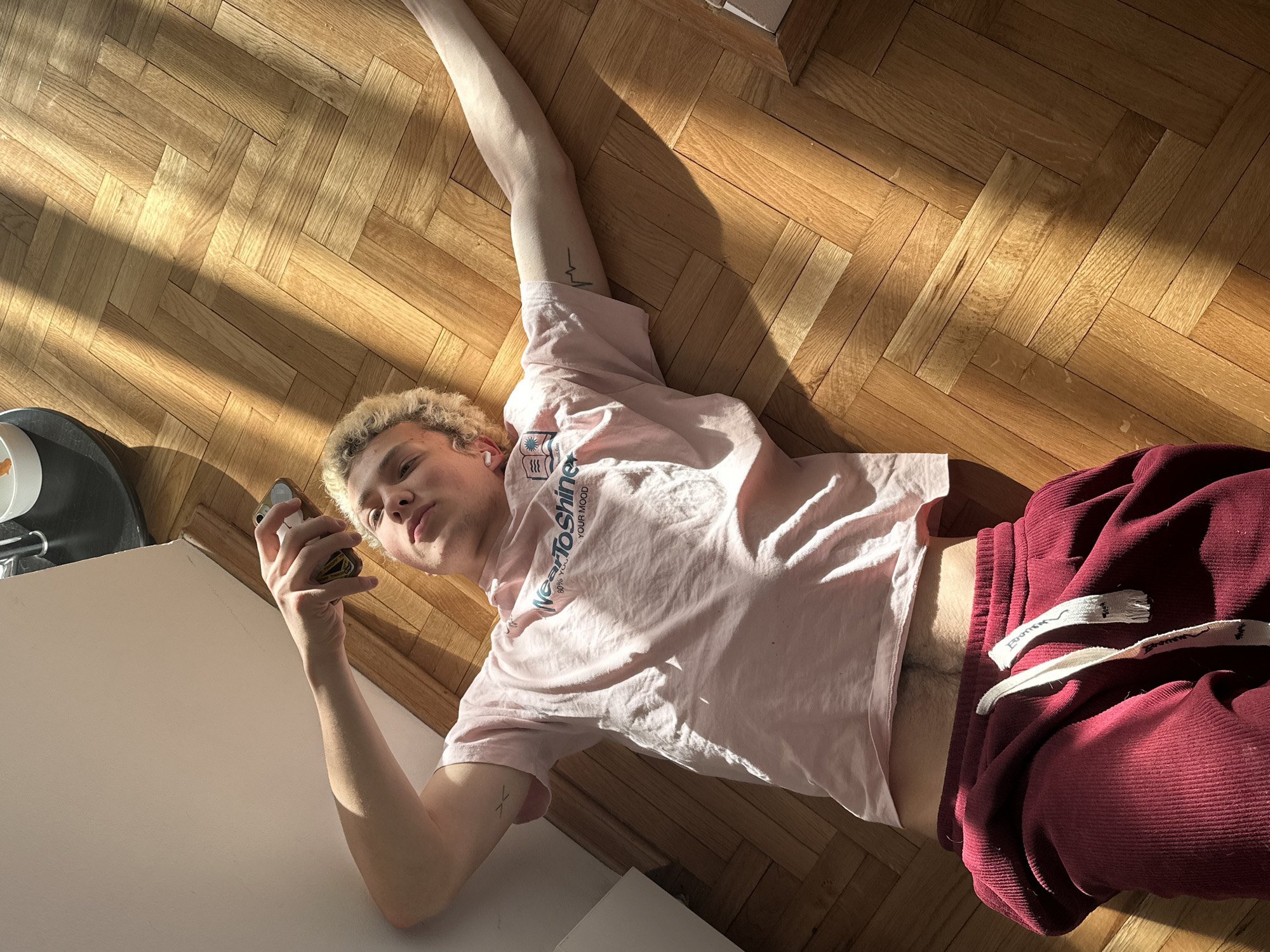 A young man with curly blonde hair lies on a herringbone wood floor, holding a cell phone in his right hand. His left arm extends straight up towards the top of the frame. He wears a light pink t-shirt with blue lettering and red corduroy shorts. He appears to be inside a room with sunlight streaming across the floor. The exact location in Novi Sad, Serbia, cannot be determined from the image.