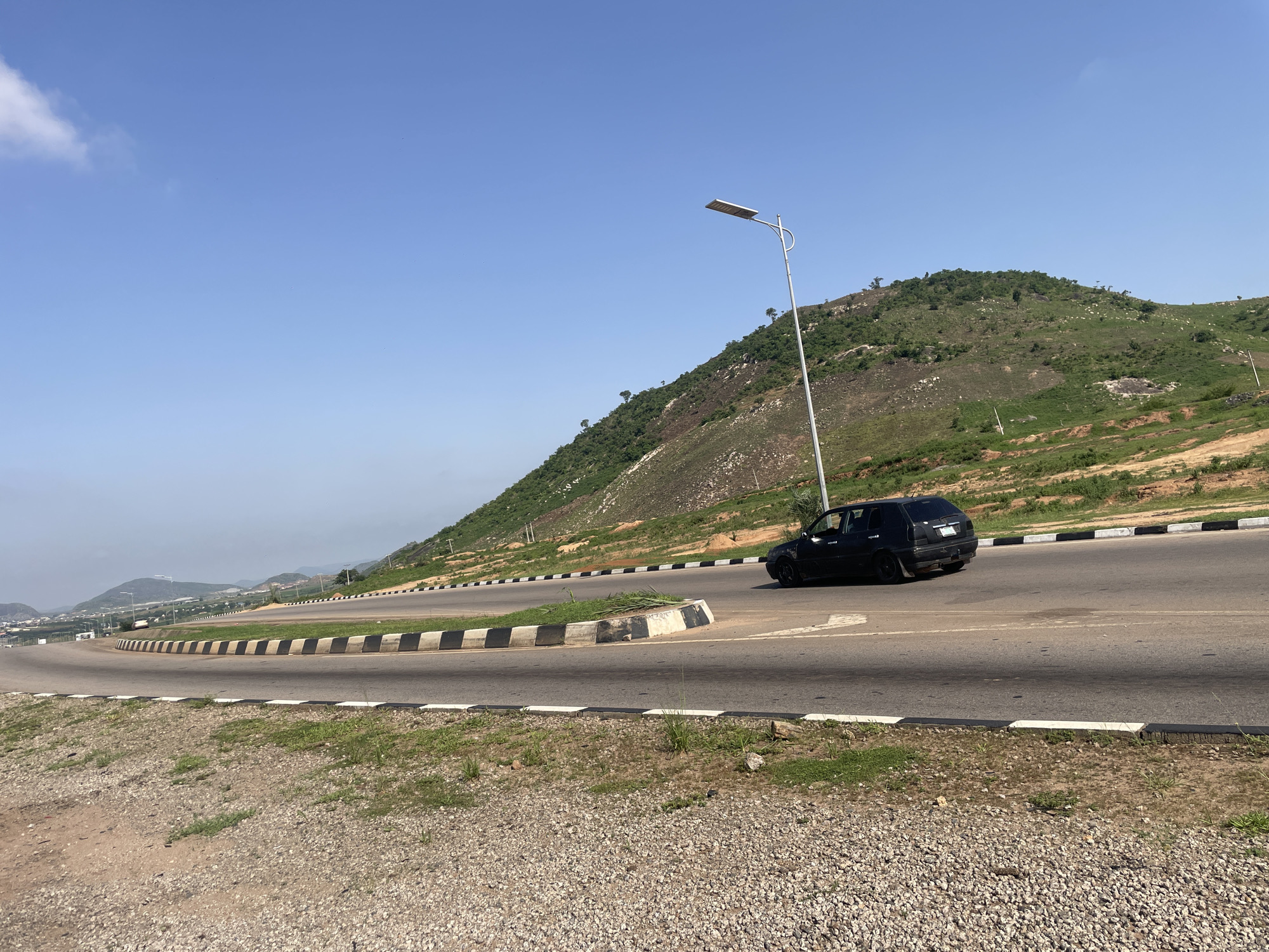 The media file captures a scenic view of a winding road in Angwan Sayawa, Nigeria. A black car is seen driving on the right side of the road, heading towards the right of the frame. The road is flanked by a concrete barrier with black and white stripes.
In the background, a lush green hill rises, dotted with trees and rocks. The sky is a clear, bright blue with a few wispy clouds. A modern streetlamp stands tall on the left side of the road, illuminating the scene. The time of day appears to be midday, with the sun casting shadows on the road and the hill.
The overall impression is one of a peaceful, natural landscape with man-made infrastructure seamlessly integrated. The location cues, such as the type of vegetation and the road design, are consistent with a tropical or sub-tropical region. There is no visible text in the media file.