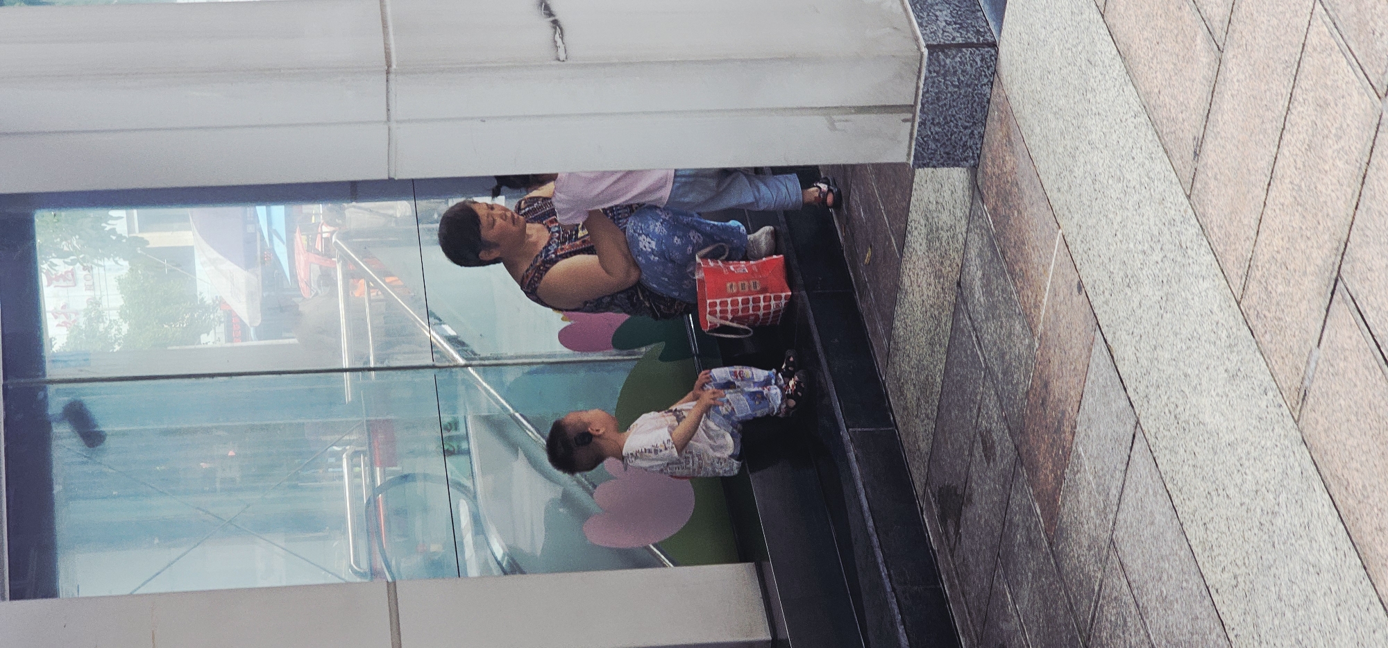 A medium-shot photograph captures a woman and a child seated on a ledge adjacent to a building's facade. The woman, positioned to the left, is wearing a patterned dress and has short, dark hair. She is next to a red shopping bag. A young child sits directly below her. The child is wearing a light-colored top and patterned pants. The setting appears to be an urban area, likely Jiulongpo District, China, based on visual characteristics. A glass facade behind the figures partially reflects a street scene, including buildings, signage, and traffic. The adjacent exterior surface is textured with stone blocks.