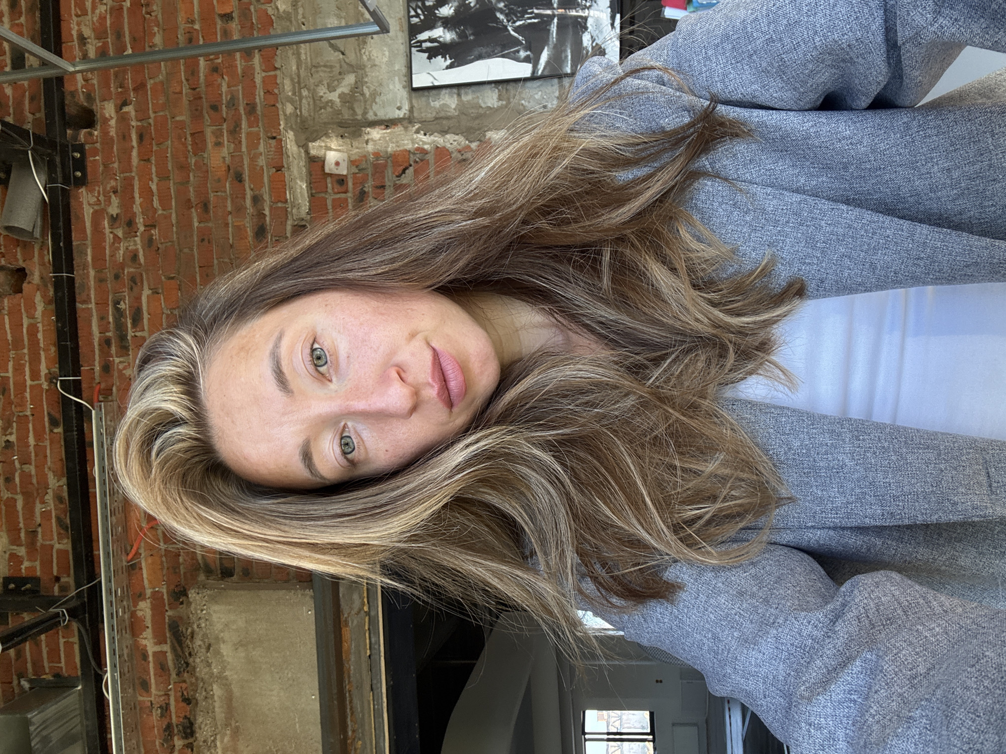 A woman is captured in a close-up, selfie-style shot, looking directly at the camera. She has long, wavy, blonde and brown hair that falls around her shoulders and a light dusting of freckles across her nose and cheeks. She is wearing a light-colored blazer over a white top. The background suggests an urban, possibly industrial or under-construction, environment with exposed brick walls, exposed pipes and wiring, and a glimpse of a window and a large black and white artwork. The scene is lit by natural light. The image does not provide any identifiable information to determine the specific location within Chaikovsky, Russia.
