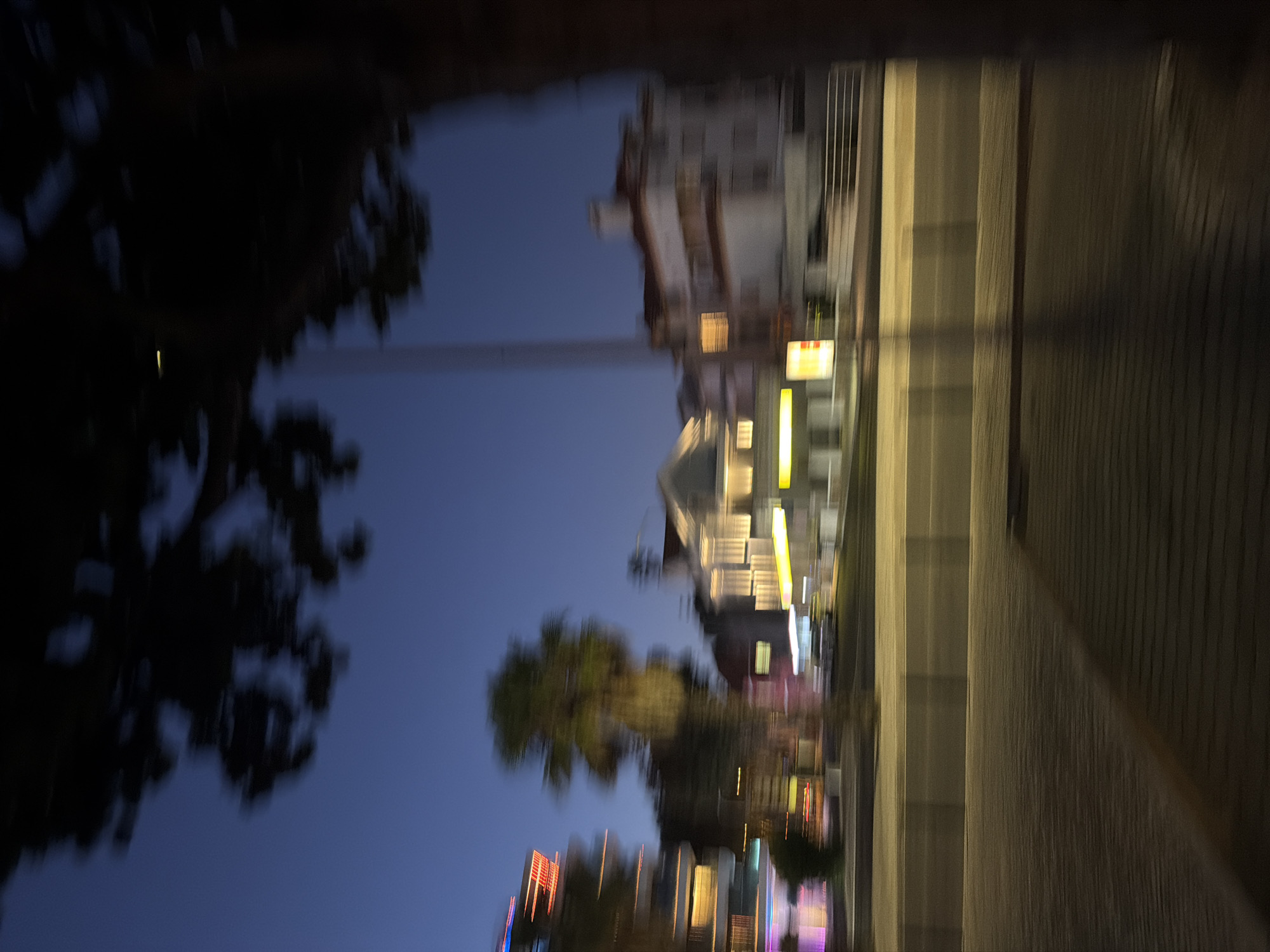 The image depicts a blurred nighttime view of a street in Cascais, Portugal. A dark tree branch obscures the top right of the frame. In the background, buildings with illuminated windows are visible against a twilight sky. To the left, a multi-story building with red and yellow lights suggests commercial activity or residences. A palm tree stands between this building and a row of white buildings with lit windows. A street with a pedestrian walkway runs through the mid-ground, with indistinct vehicles possibly present. The overall impression is one of motion blur, possibly from a moving vehicle, capturing the ambiance of a European coastal town at dusk.