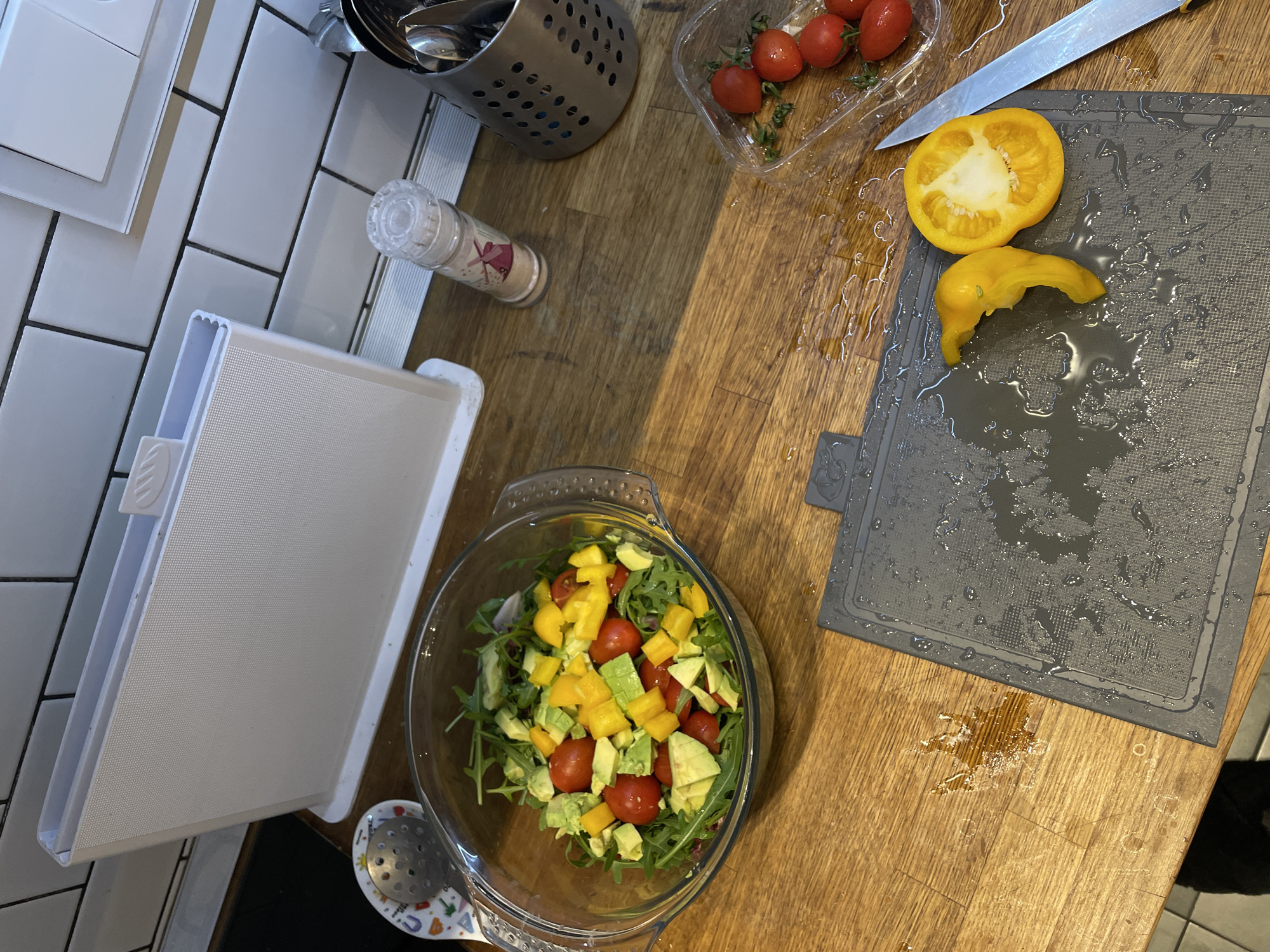 The image shows a kitchen counter with various items related to food preparation. There's a salad in a glass bowl containing tomatoes, yellow peppers, and greens. Next to it, a white container is placed near a tiled wall. On the wooden counter, a utensil holder, a spice container, and a plastic container with tomatoes can be seen. A grey cutting board with a sliced yellow pepper and a knife lies on the counter, with water droplets scattered around. The scene suggests a salad preparation process. The location, Moscow, Russia, is not apparent from the image's content.