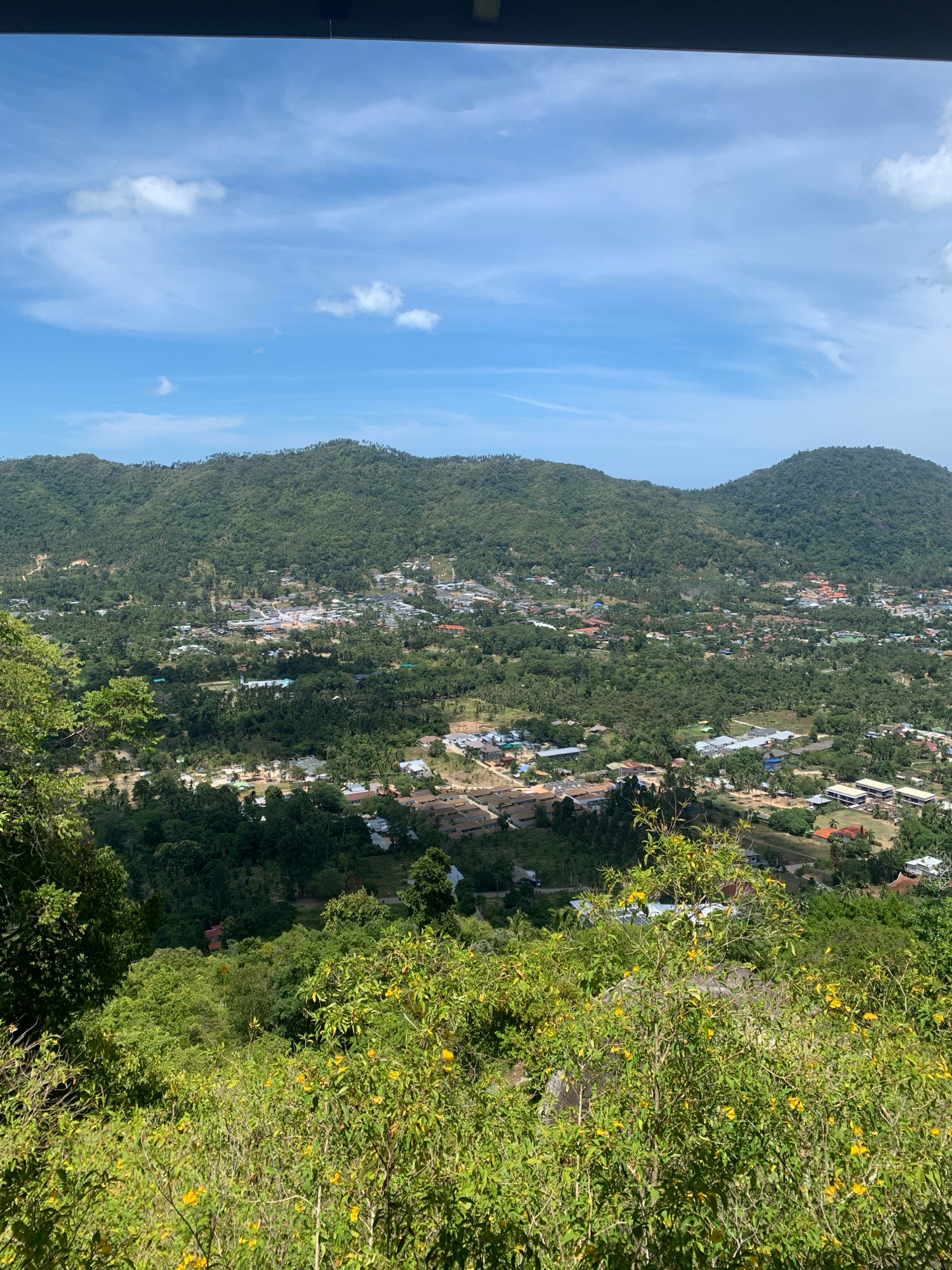 A high-angle, wide shot captures a verdant hillside overlooking a coastal town in Ko Samui District, Thailand. The foreground is dominated by dense green foliage, interspersed with bright yellow flowers. Beyond this, the midground reveals a sprawl of buildings, appearing to be homes and small businesses, nestled amongst the trees and rolling hills. The structures are a mix of white and terracotta-roofed buildings, some with visible solar panels. The background is defined by lush, tree-covered mountains that recede into the distance under a bright blue sky dotted with wispy white clouds. The overall impression is one of a tropical landscape that has been partially developed for settlement.