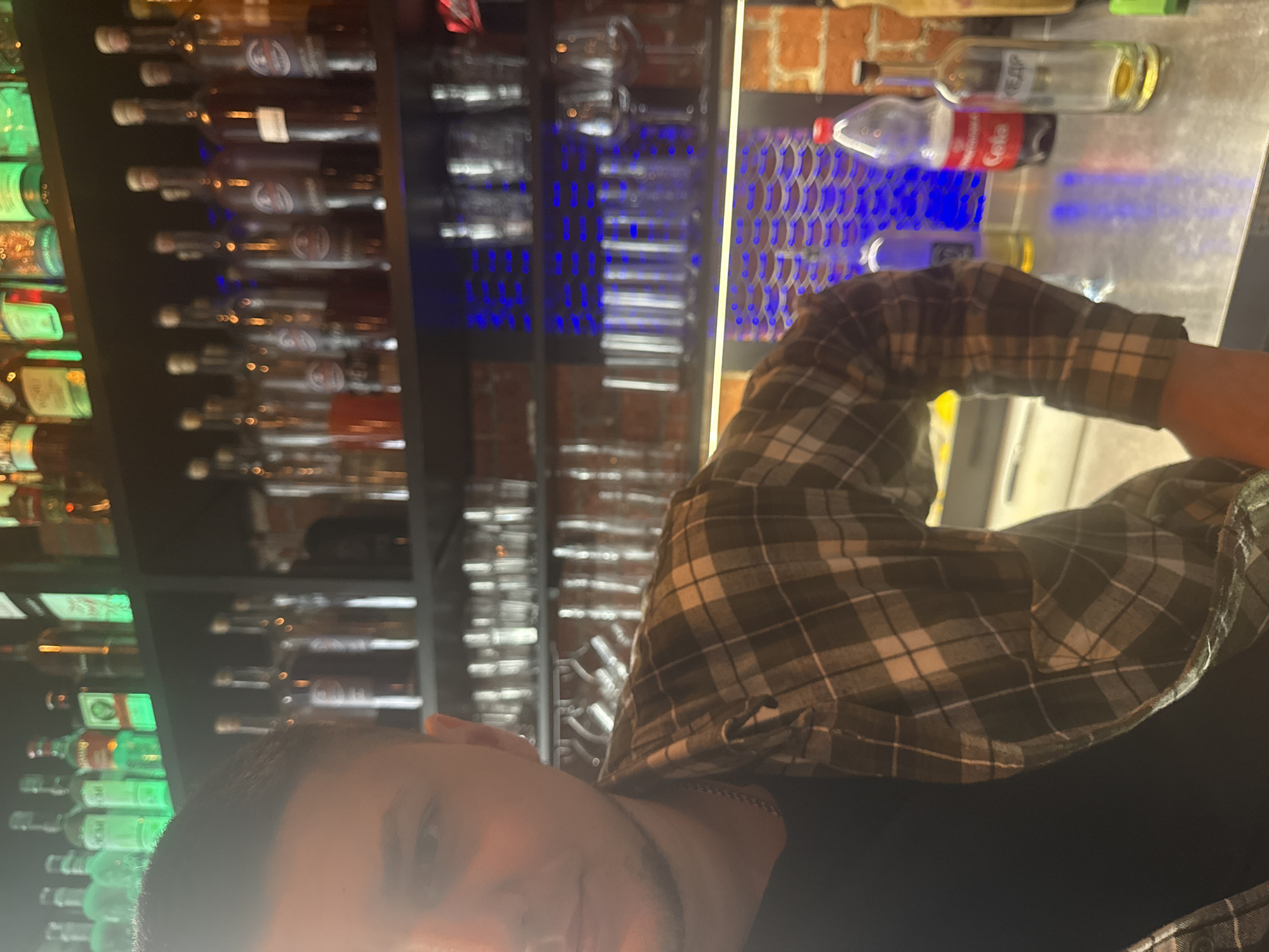 The photograph depicts a bar setting. In the foreground, part of a person is visible, wearing a black shirt and a checkered flannel shirt. Behind the person, shelves hold numerous bottles, possibly liquor. A bar counter can be seen, featuring bottles of Coca-Cola and liquor. Above the counter, there's a brick wall and a decorative panel with blue lights. The location may be a bar in Moscow, Russia, but this is not confirmed by the visible content.