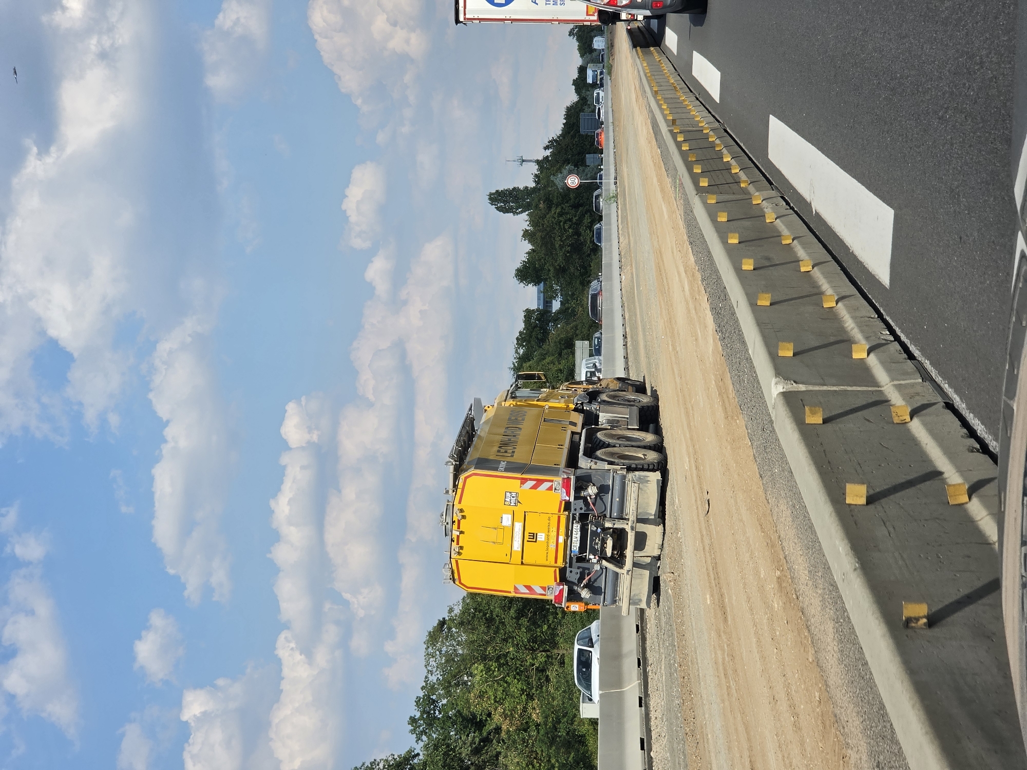 The image shows a yellow construction vehicle with "LEONHARD WEISS" written on its side, operating on a highway under construction in Rüsselsheim am Main, Germany. The highway is undergoing road work, with a new lane being constructed. The vehicle appears to be a water truck or a similar piece of heavy machinery used for road construction. In the background, there are trees and a cloudy sky. The overall scene depicts an active construction site on a highway.