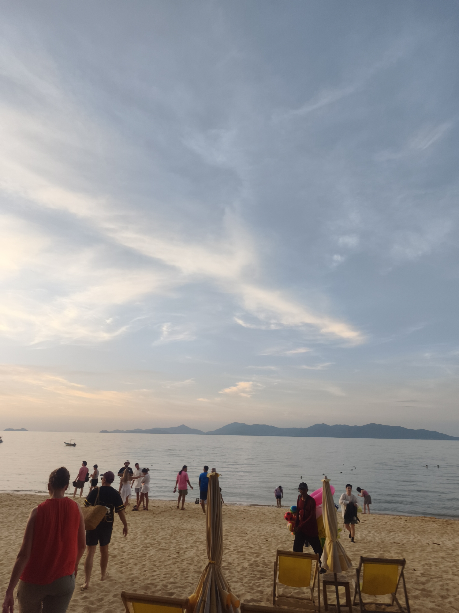 In Ko Samui District, Thailand, a beach scene unfolds with a gentle sky as the backdrop. The sky is a soft blend of blues and whites, with streaks of pale orange and pink near the horizon, suggesting either sunrise or sunset. A body of water, likely the sea, stretches out to meet a distant mountain range. The water is calm, with a few small boats visible offshore.
On the sandy beach, people are dispersed, some walking, others gathered in small groups. A woman in a red top is in the foreground, facing away from the camera. To her left, a man in a dark shirt and shorts carries a straw bag. Other individuals are seen in the middle ground, some in brightly colored clothing. Among them, a person dressed in red appears to be a vendor, holding a cluster of colorful balloons. Beach chairs, some with umbrellas, are positioned on the sand. The overall impression is one of a relaxed, late afternoon or early morning atmosphere on a tropical beach.