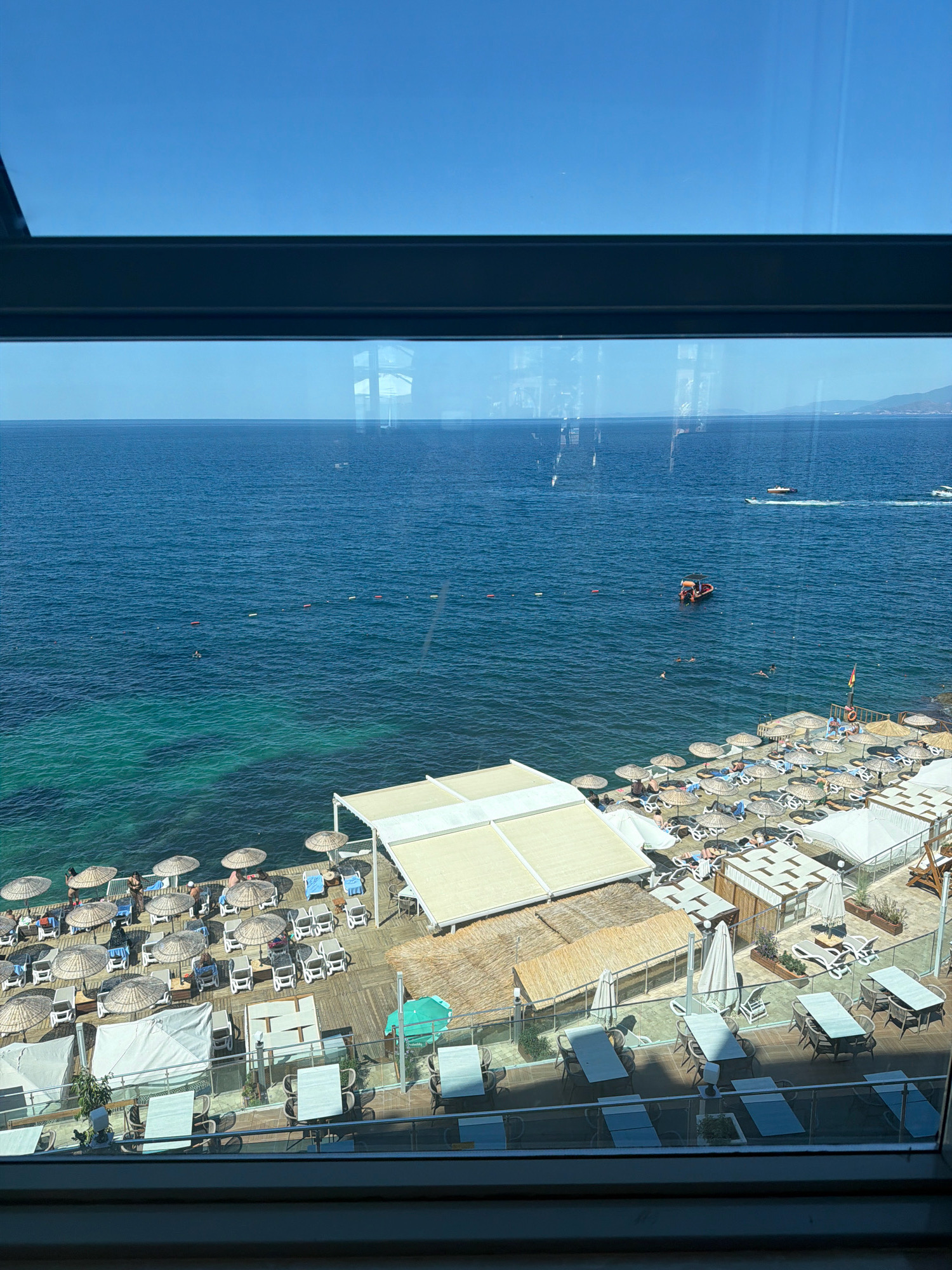 This media file captures a vibrant beach scene from an elevated perspective, likely a hotel room or balcony overlooking the Aegean Sea in Kuşadası, Turkey. The image depicts a sun-drenched day with a clear blue sky and calm, turquoise waters.
The foreground features a meticulously arranged beach resort area with numerous thatched umbrellas providing shade over rows of lounge chairs. Several people are scattered amongst the loungers, some appearing to be relaxing, while others may be swimming or wading in the shallow, clear water near the shore. A wooden pier extends into the sea, also lined with umbrellas and chairs. Beyond the immediate beach area, the expansive blue sea stretches to the horizon, dotted with a few small boats and a distant coastline.
In the water, several individuals can be seen swimming, and a small orange boat with people on board is anchored or moving slowly. A few buoys mark a designated swimming area. The scene conveys a sense of leisure and relaxation, typical of a summer vacation destination. The presence of multiple shaded seating areas, the calm sea, and the overall arrangement of the resort suggest it is a popular spot for tourists seeking sun and sea. The lighting indicates it is likely midday or early afternoon, with the sun high in the sky casting distinct shadows. No visible text is present in the image.