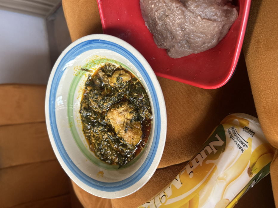 A meal is presented indoors in Egbe, Nigeria. The scene features a white ceramic bowl with blue and green striped rim, containing a dark green leafy soup or stew with several pieces of cooked poultry and reddish oil. To the right, a rectangular red plastic plate holds a single lump of brownish-purple swallow food, consistent with amala or fufu. A 500ml "NutriMilk" banana-flavored milk drink in a yellow and white flexible package is positioned to the lower left, displaying text "Nutritious milk with natural banana taste!". All items rest on a textured, orange-brown upholstered surface, likely a sofa or chair, with a similar upholstered surface and a light-colored wall visible in the background. No individuals are present.