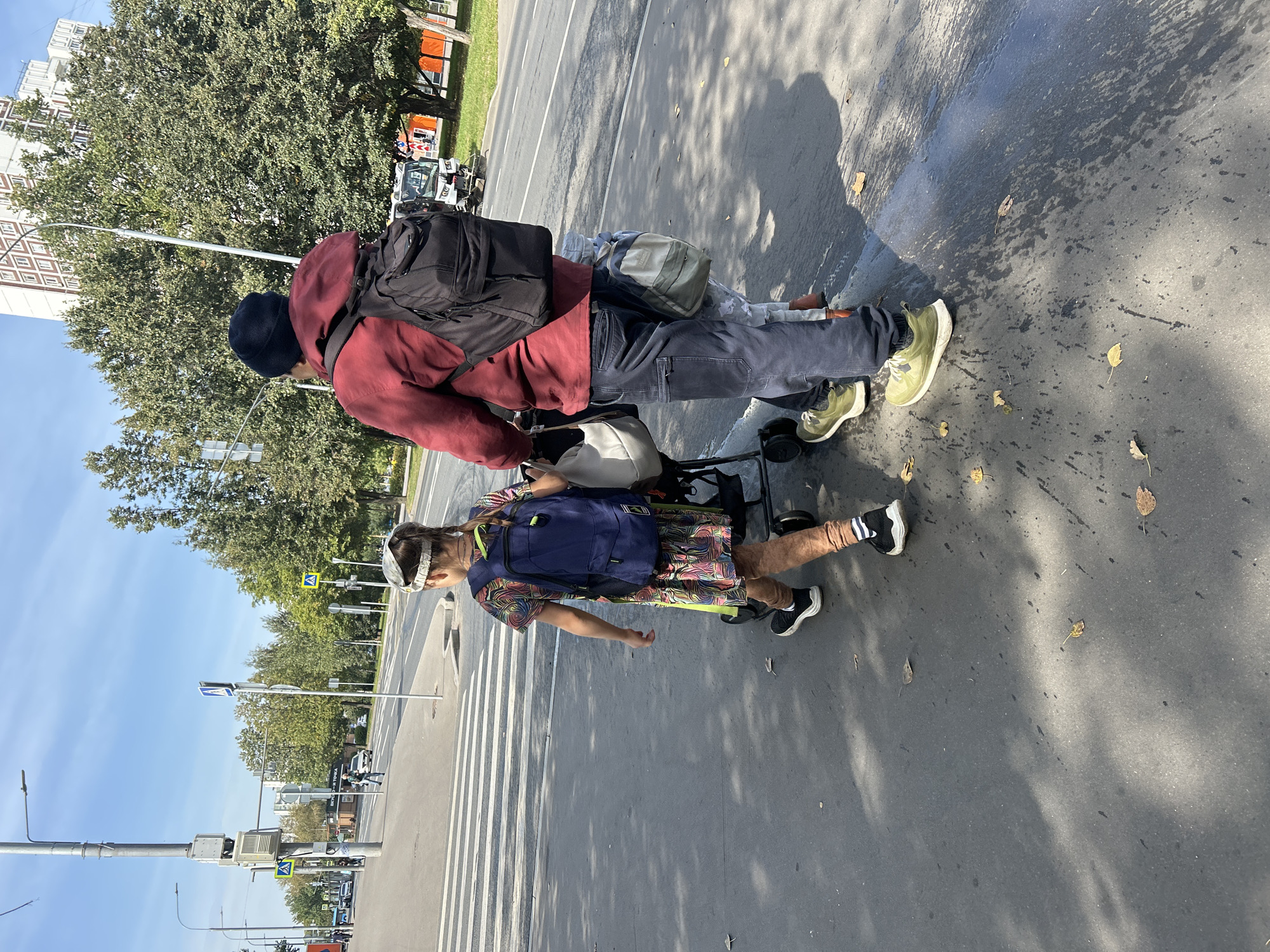 The image depicts a street scene in Moscow, Russia, on a sunny day. A family, consisting of at least two adults and one child, is walking across a pedestrian crossing. The child, a young girl, walks slightly ahead, pulling a stroller. She is wearing a colorful dress, brown leggings, and black sneakers, and has a blue backpack. Behind her, an adult wearing a red hooded jacket, dark pants, and a black beanie walks while holding onto the stroller. This adult also has a black backpack. Another adult, mostly obscured, is walking behind the person in the red jacket, carrying a gray and beige backpack. The background shows a wide street with cars, trees, buildings, and streetlights. The pedestrian crossing is marked with white stripes, and there are fallen leaves on the asphalt.