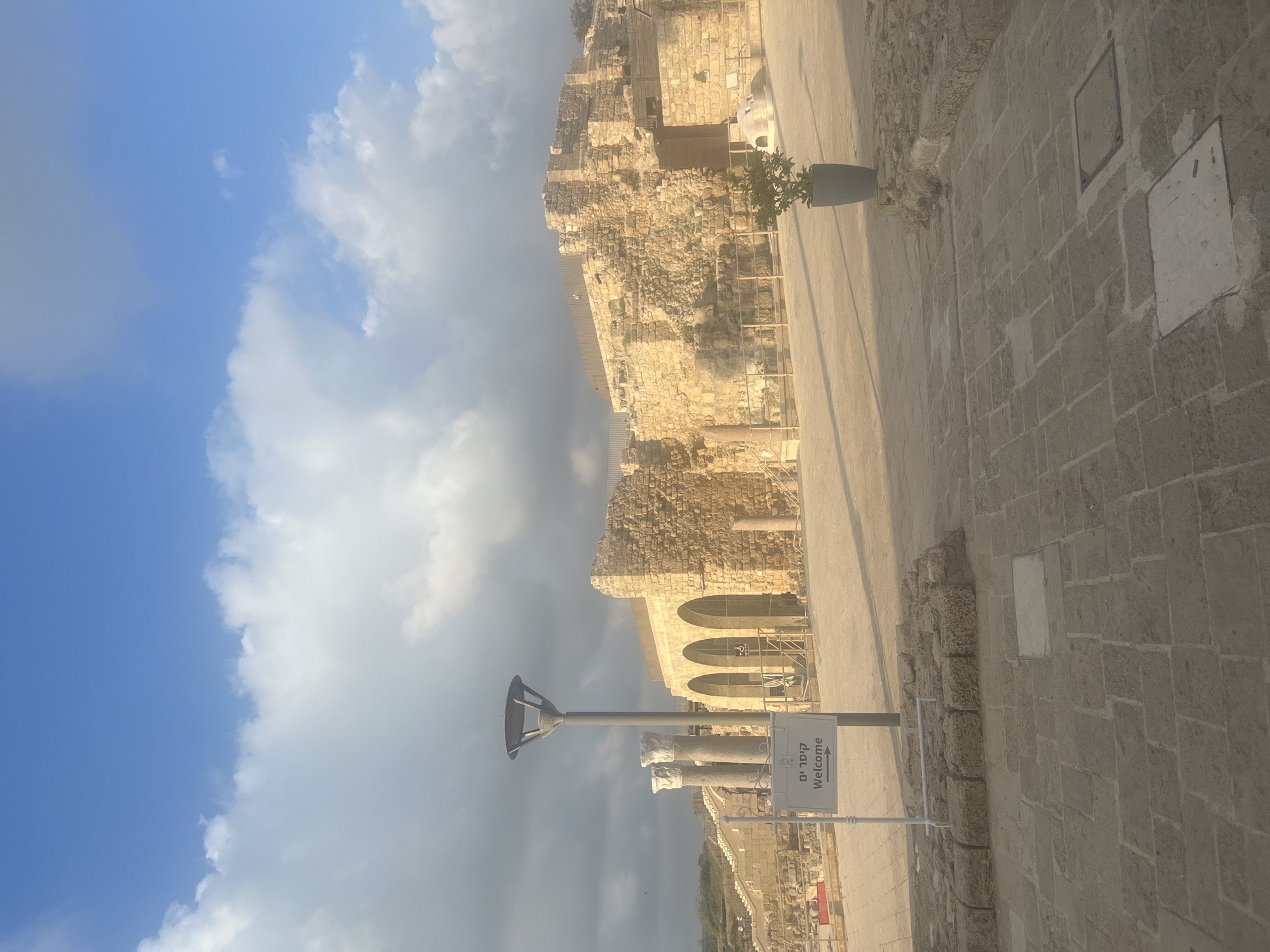 The image depicts a sunny day at an archaeological site in the city of Hof HaCarmel Regional Council, Israel. In the foreground, a white sign with "Welcome" written in English and Hebrew stands beside a lamppost. The ground is paved with large stone tiles. Beyond the sign, ancient stone structures rise, featuring arches and weathered walls. In the mid-ground, there are rows of stone columns. To the right, a potted plant sits near the stone ruins. The sky above is a bright blue with scattered clouds.