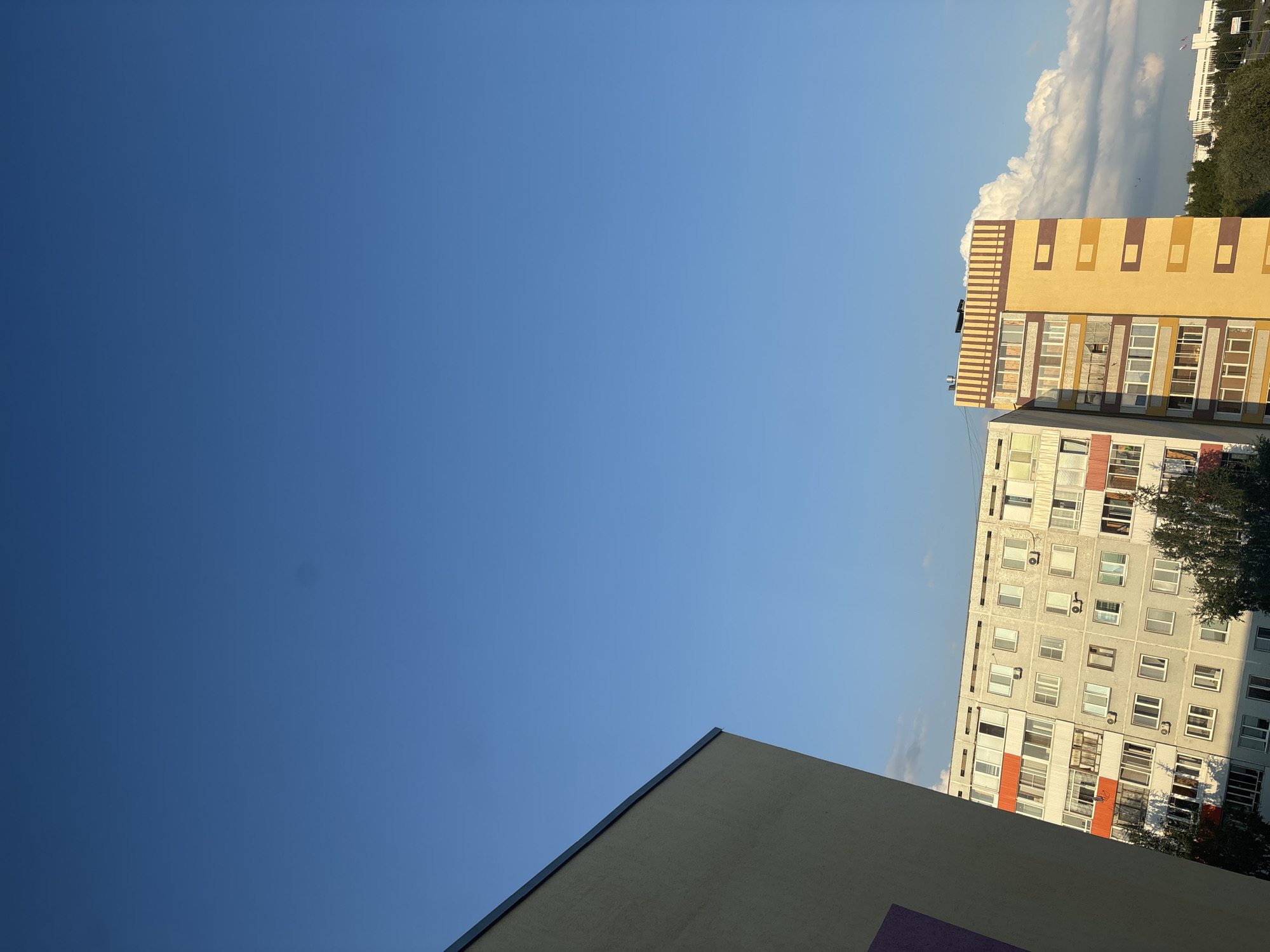 The image captures a view of residential buildings against a clear blue sky with some scattered clouds in the distance. On the left, a light-colored building with a dark trim line diagonally cuts across the frame. To the right of this, two apartment buildings are visible. The first apartment building is multi-story and appears to be constructed of gray concrete with numerous windows. It has sections with orange balconies. Next to it, a taller, narrower building is painted in yellow and brown hues, also featuring multiple windows and balconies. The buildings are likely situated in a residential area of Naberezhnye Chelny, Russia, given the architectural style commonly found in Russian cities. Trees are visible at the base of the buildings and in the background, where a wider, more modern-looking building with a flat roof and a row of flagpoles is also present. The lighting suggests it is late afternoon or early evening, with the sun casting shadows. No people are discernible in the image.