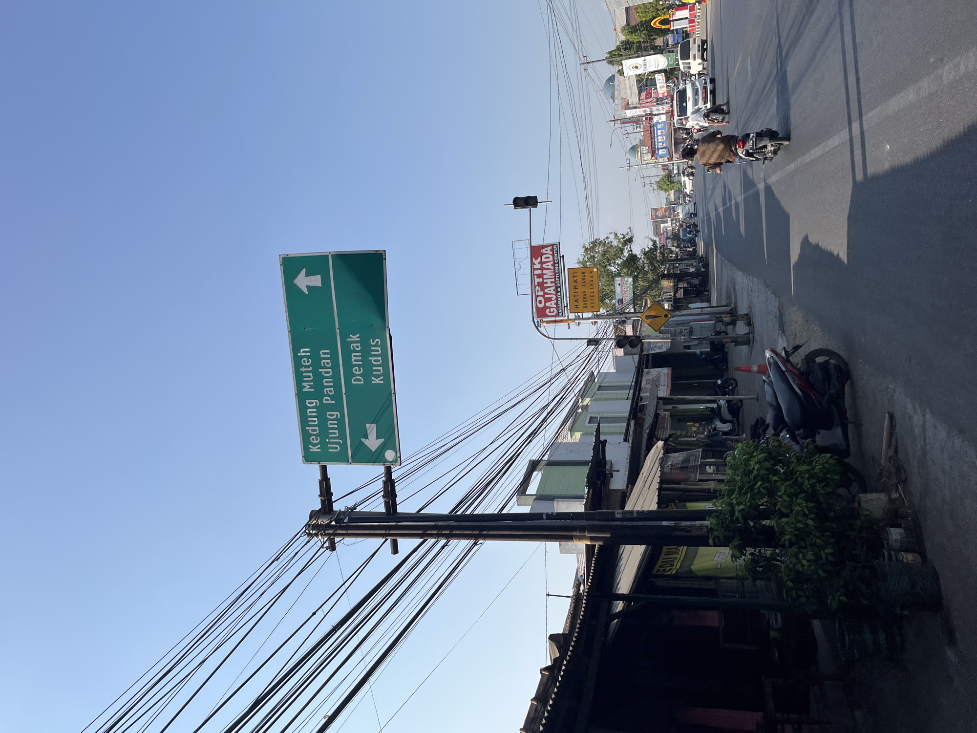 This is a daytime, eye-level, medium shot of a street scene in Kalinyamatan, Indonesia, captured on a clear, sunny day. The sky is a vibrant blue, with no clouds visible. A traffic light hangs above the road, and a signpost with directional arrows indicates routes to "Kedung Muteh Ujung Pandan" and "Demak Kudus." The sign for "Kedung Muteh Ujung Pandan" has an upward-pointing arrow, while the sign for "Demak Kudus" has a left-pointing arrow. To the left of the road, there are several shops with awnings and a dense green bush in a pot. A dark-colored motorcycle with a red stripe is parked near the bush. On the right side of the road, multiple vehicles, including cars and motorcycles, are traveling. One motorcycle rider, wearing a brown shirt and dark pants, is in the foreground, facing away from the camera and riding down the street. Further down the road, white cars and other motorcycles are visible, along with more shops and buildings lining the street. A traffic light with red, yellow, and green signals is positioned at an intersection. Another sign for "Optik Gajahmada" and "Hati-hati Daerah Rawan Kecelakaan" (Caution: Accident-prone area) is also present. Power lines are strung across the frame, supported by utility poles, creating a complex network of wires. The road has white lane markings. The overall impression is of a bustling urban street.