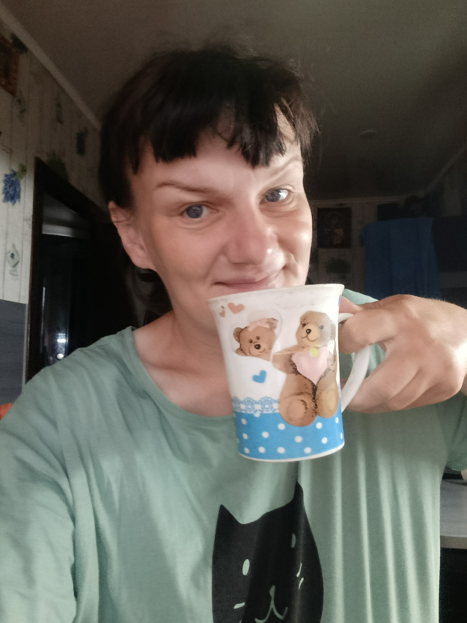 A woman is holding a mug with two teddy bears on it, one of which is wearing a heart-shaped bib. The mug has a white handle and a blue section with white polka dots at the bottom. The woman has short brown hair with bangs and blue eyes. She is wearing a green t-shirt with a black cat face printed on it. The background shows a room with light-colored patterned wallpaper and a doorway or opening. A blurry picture and a blue garment hang on the wall. The location of Фоки, Russia is unlikely, as this information cannot be determined from the image alone.