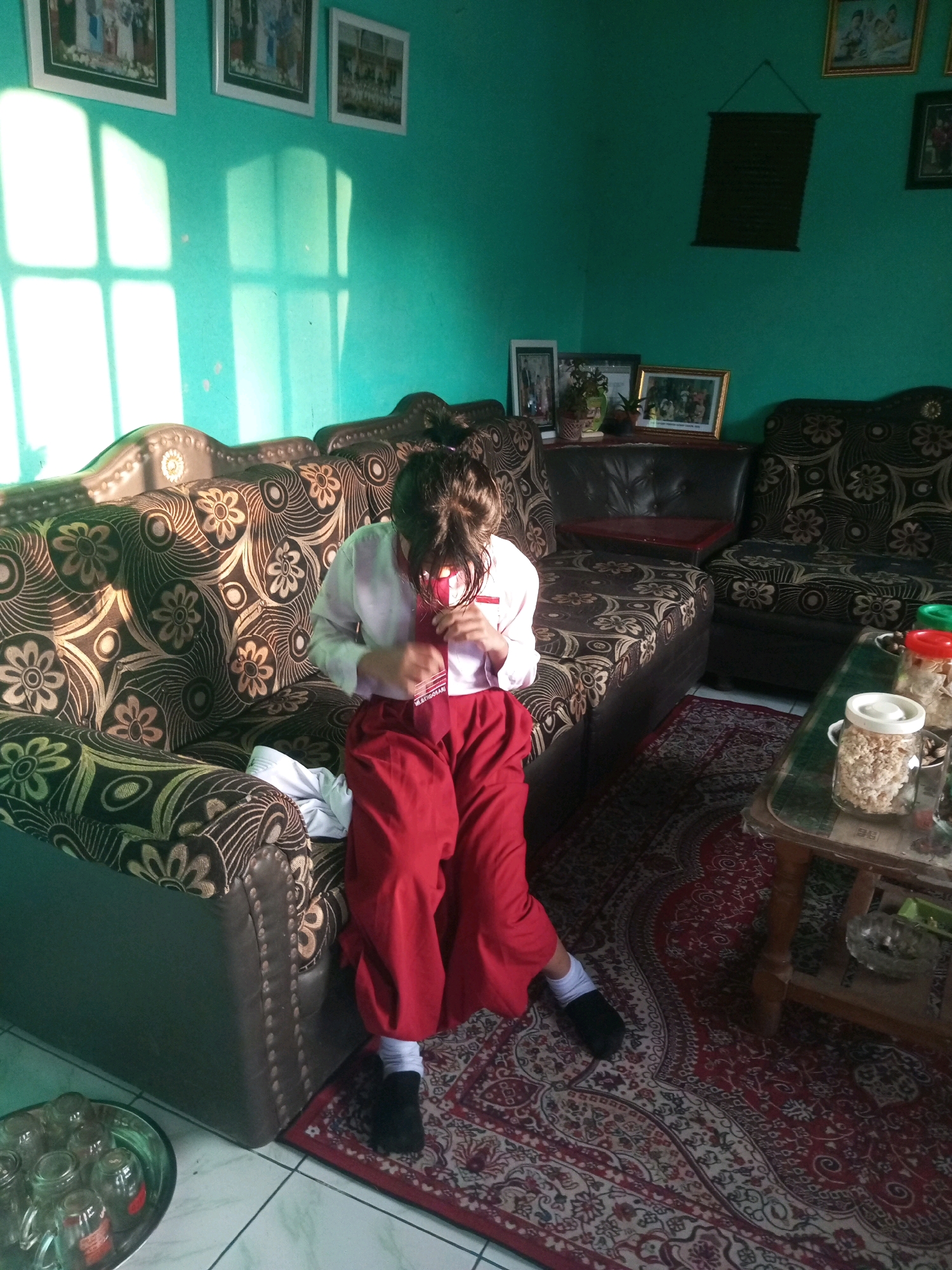 The image depicts a girl in a school uniform, consisting of a white shirt and red pants, sitting on a patterned couch in what appears to be a living room. She is adjusting something near her neck, possibly a tie or scarf. The room has a teal-colored wall with framed pictures hanging on it, and sunlight is streaming through a window, casting geometric patterns on the wall. A patterned rug covers part of the floor, and a wooden coffee table with containers of snacks is visible to the right. The scene suggests a domestic setting in Karangsari, Indonesia, possibly a school break or after-school moment.