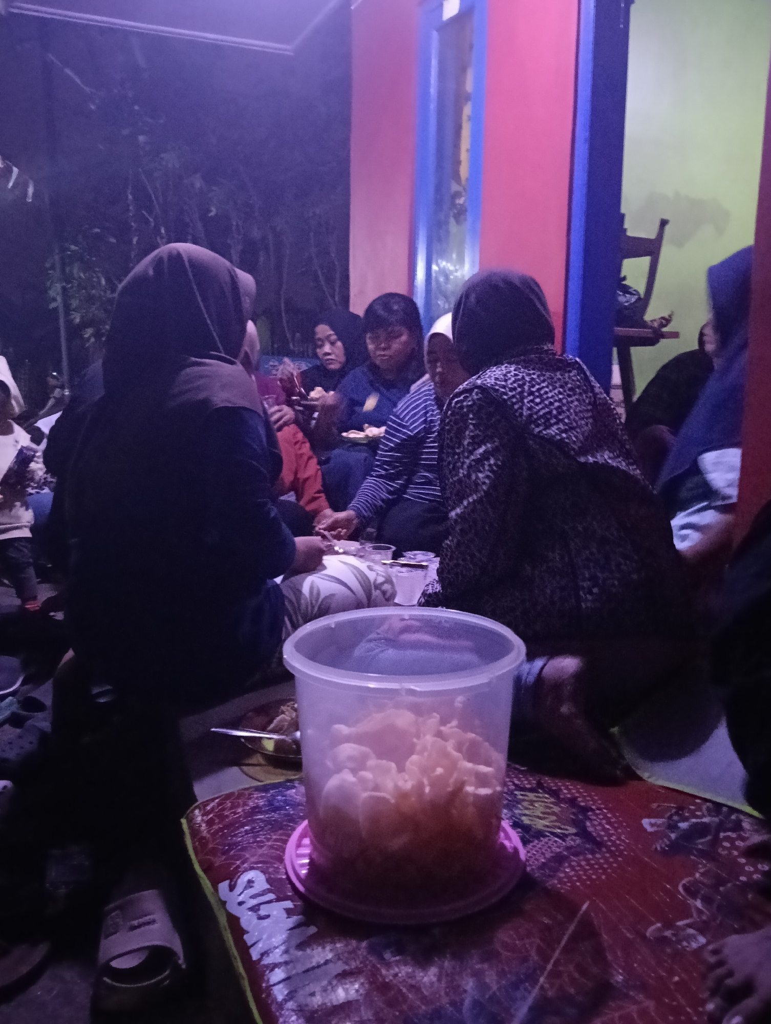 The image depicts a gathering of people, predominantly women, seated on the floor in what appears to be a residential area at dusk or night. The setting is dimly lit, with purple and pink light dominating the scene, suggesting artificial lighting. The people are mostly facing away from the camera, engaged in conversation and possibly eating. One woman in the foreground is wearing a black hijab and a patterned top. A clear plastic container filled with light-colored snacks, likely crackers or crisps, is prominent in the lower center of the image. The ground is covered with mats or blankets, one of which has a colorful, possibly Indonesian, pattern with stylized writing. In the background, a doorway with a blue frame and pink and green walls is visible, along with hints of foliage outside. The presence of people sitting on the floor and the casual attire suggest a relaxed, informal gathering. The mention of Tasikmalaya, Indonesia, implies the cultural context of the gathering, likely a local tradition or social event.