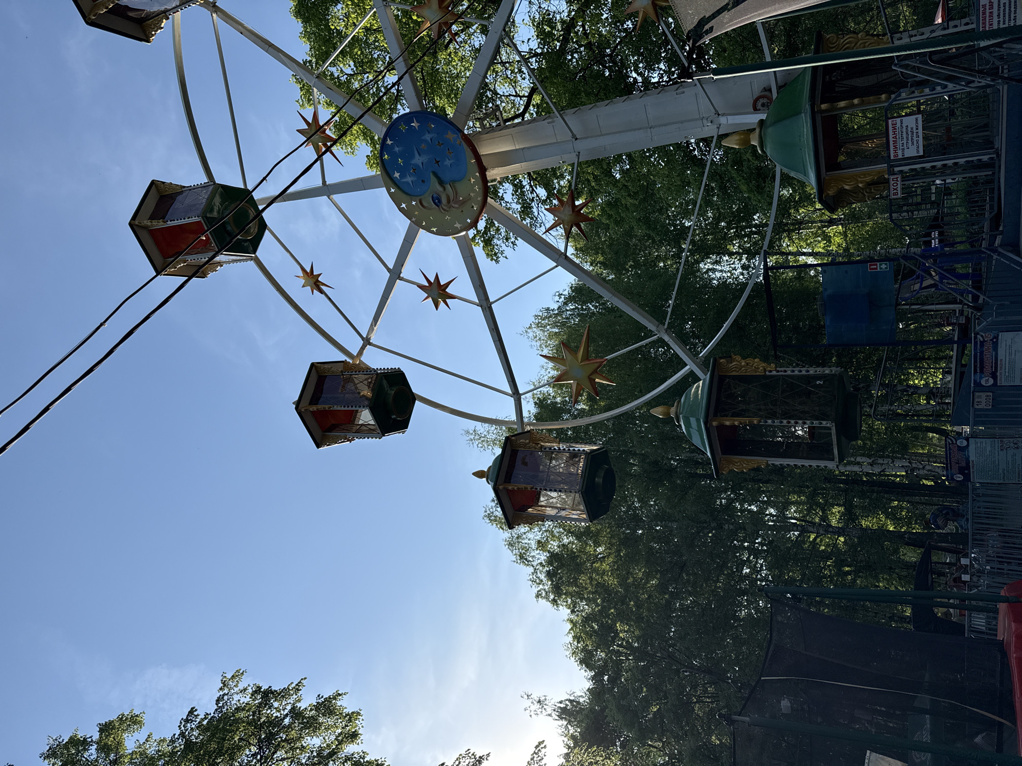 The image depicts a small, antiquated Ferris wheel in a park setting, located in Izhevsk, Russia. The Ferris wheel features enclosed cabins, painted in various colors. Each cabin has a distinctive shape with multiple windows. Decorative stars are affixed to the wheel's structure. The central hub of the wheel displays a celestial motif with a moon and stars.
The sky is blue with some high clouds visible near the sun, implying it is daytime. Tall trees surround the Ferris wheel, giving the impression of being in a wooded or park area. Signs with Russian text can be seen, suggesting the geographical location. There are no visible people.