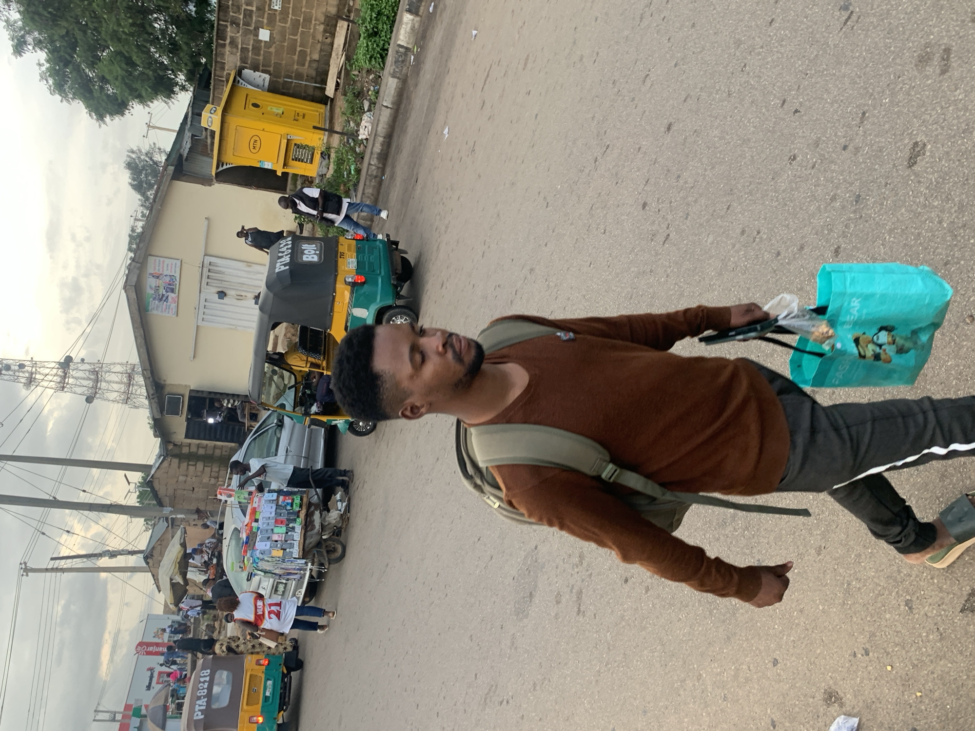 In Barakin Lerawi, Nigeria, a man with a backpack walks across a paved street, holding a turquoise shopping bag and a plastic bag containing food items. He wears a brown long-sleeved shirt and dark pants with a white stripe down the side. In the background, several auto-rickshaws and a silver car are parked along the roadside. A vendor stands beside a cart laden with mobile phones, while other pedestrians and shops are visible further down the street. A yellow MTN booth stands near a brick wall, and a large cell tower is visible in the distance. The sky is overcast.