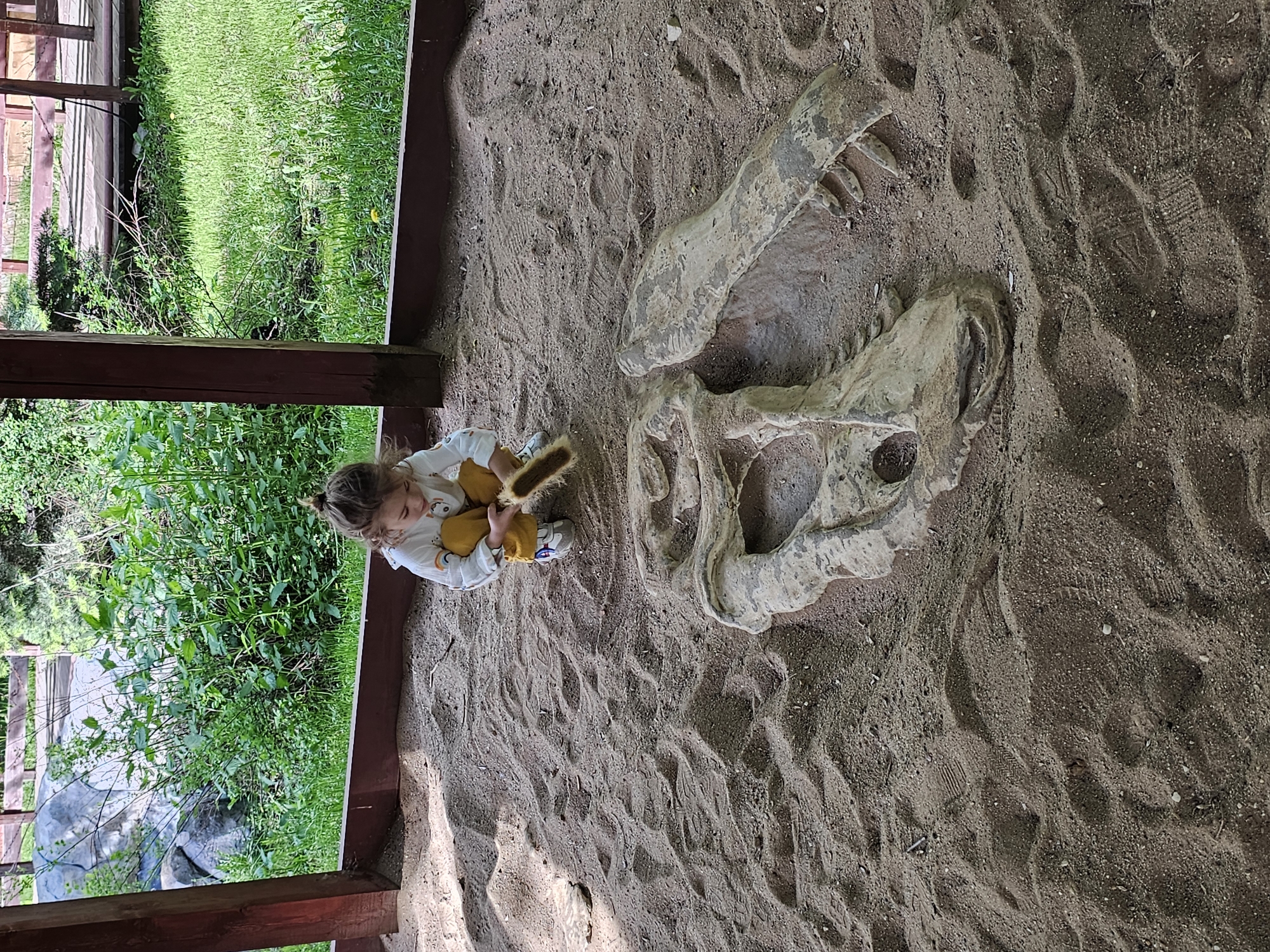 A young child is seated in a sandpit next to a large dinosaur skull replica. The sandpit is under a wooden structure with green vegetation visible beyond the frame. There is no evidence in the image to suggest this photo was taken in Khimki, Russia.