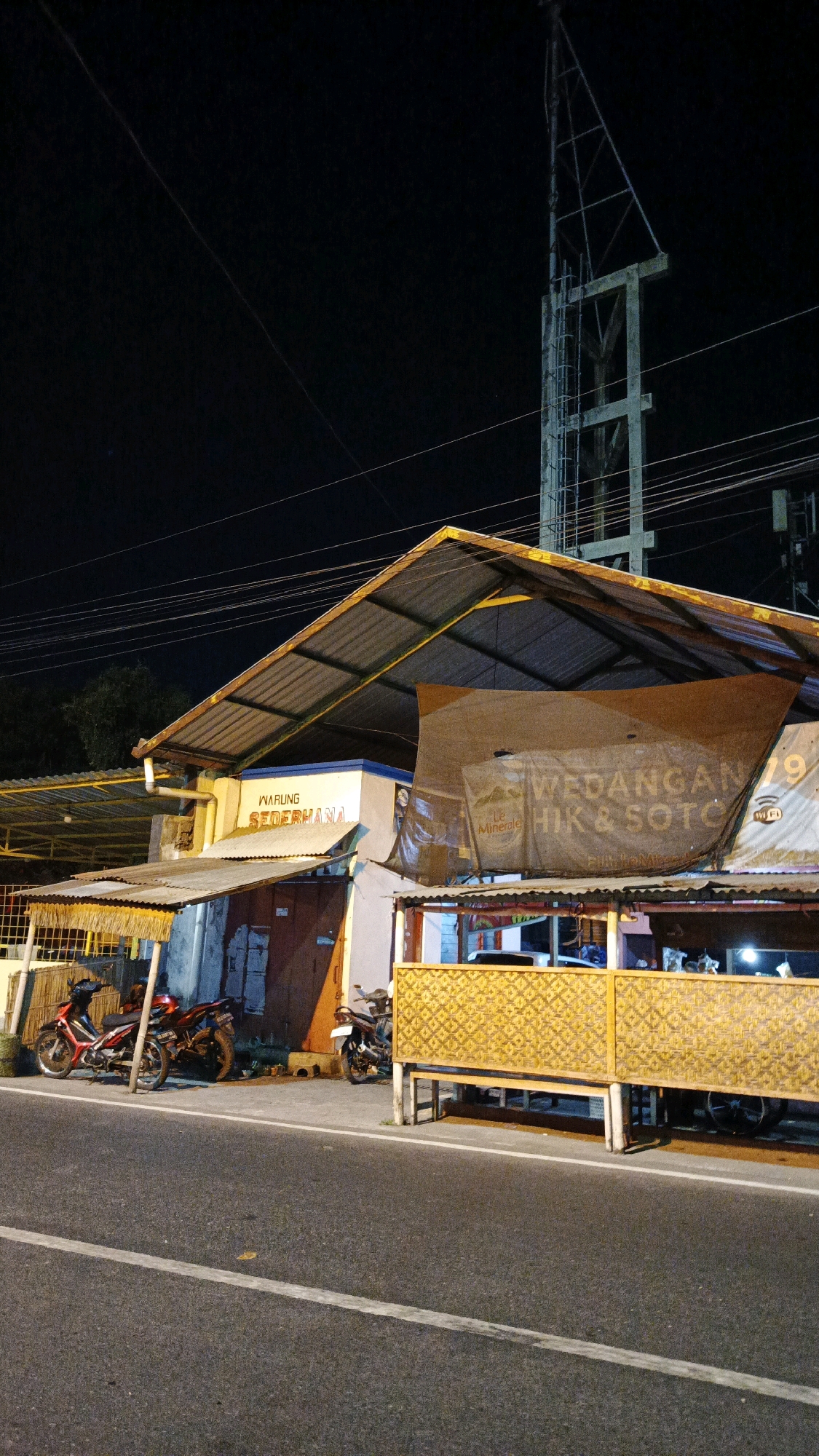 The image depicts a roadside scene at night in Surakarta, Indonesia. A modest establishment, identified by signage as "WARUNG SEDERHANA" and "WEDANGAN HIK & SOTO," is visible. The building has a metal-sheeted roof that extends over a seating area. The facade features signage including "Le Minerale" and a Wi-Fi symbol. In front of the establishment, parked along the roadside, are two motorcycles. One is a red and black motorcycle parked to the left, and another dark-colored motorcycle is parked to the right. The road surface is asphalt with white lane markings. In the background, a tall communication tower is visible against the dark night sky, along with electrical wires. The lighting suggests artificial illumination from the establishment and possibly streetlights.
