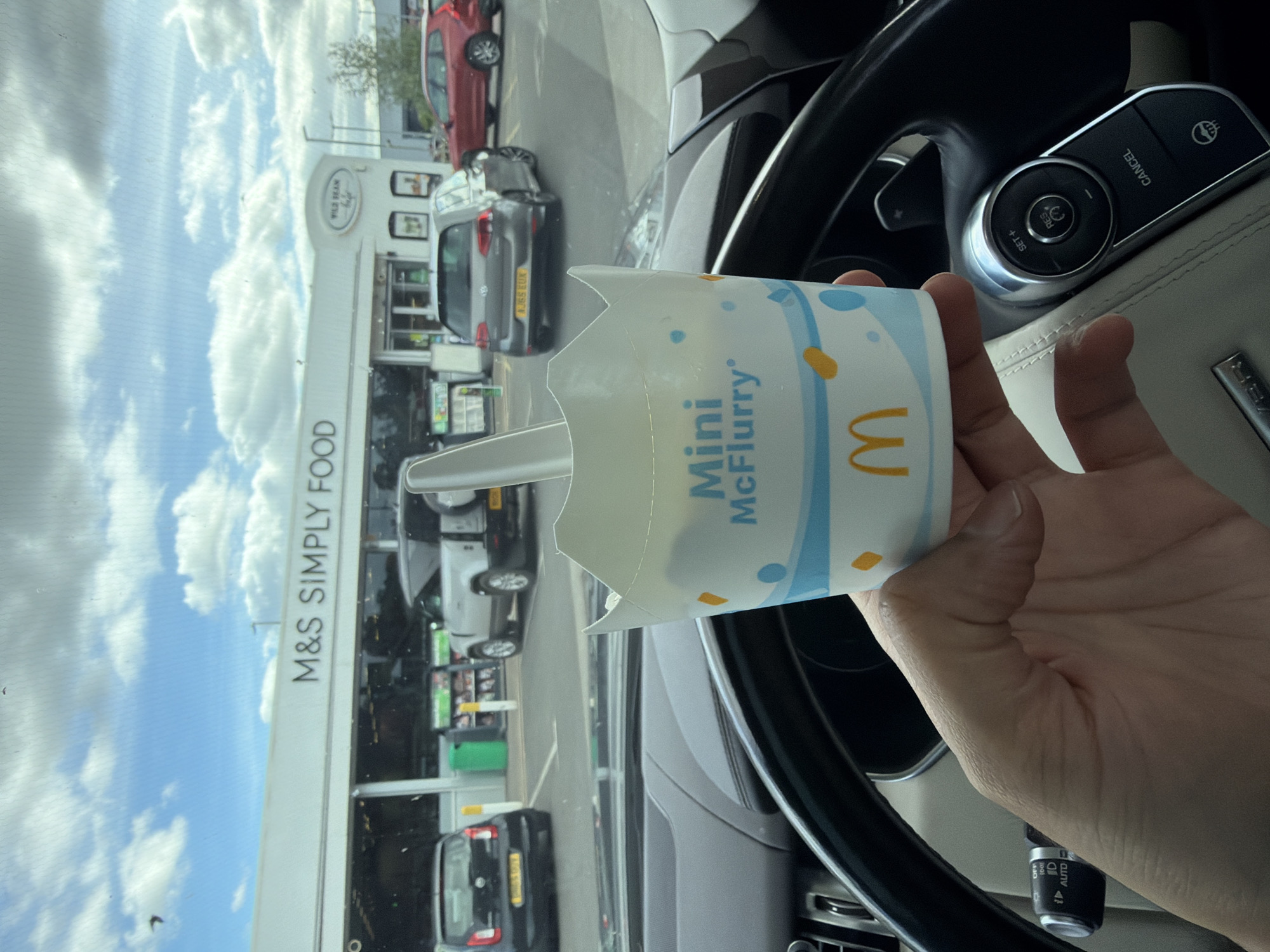 A person is holding a Mini McFlurry cup from McDonald's inside a car. The car is parked in front of an M&S Simply Food store, identified by the signage on the building. The background shows several cars parked in a lot, and a red car is visible on the right. The sky is cloudy with patches of blue. The M&S Simply Food is located at a petrol station, as indicated by the presence of fuel pumps and a "Wild Bean Cafe" sign. The scene appears to be a typical roadside convenience store and cafe. The location can be identified as the M&S Simply Food at the BP petrol station on Newport Road, Caldicot, NP26 5FU, United Kingdom.