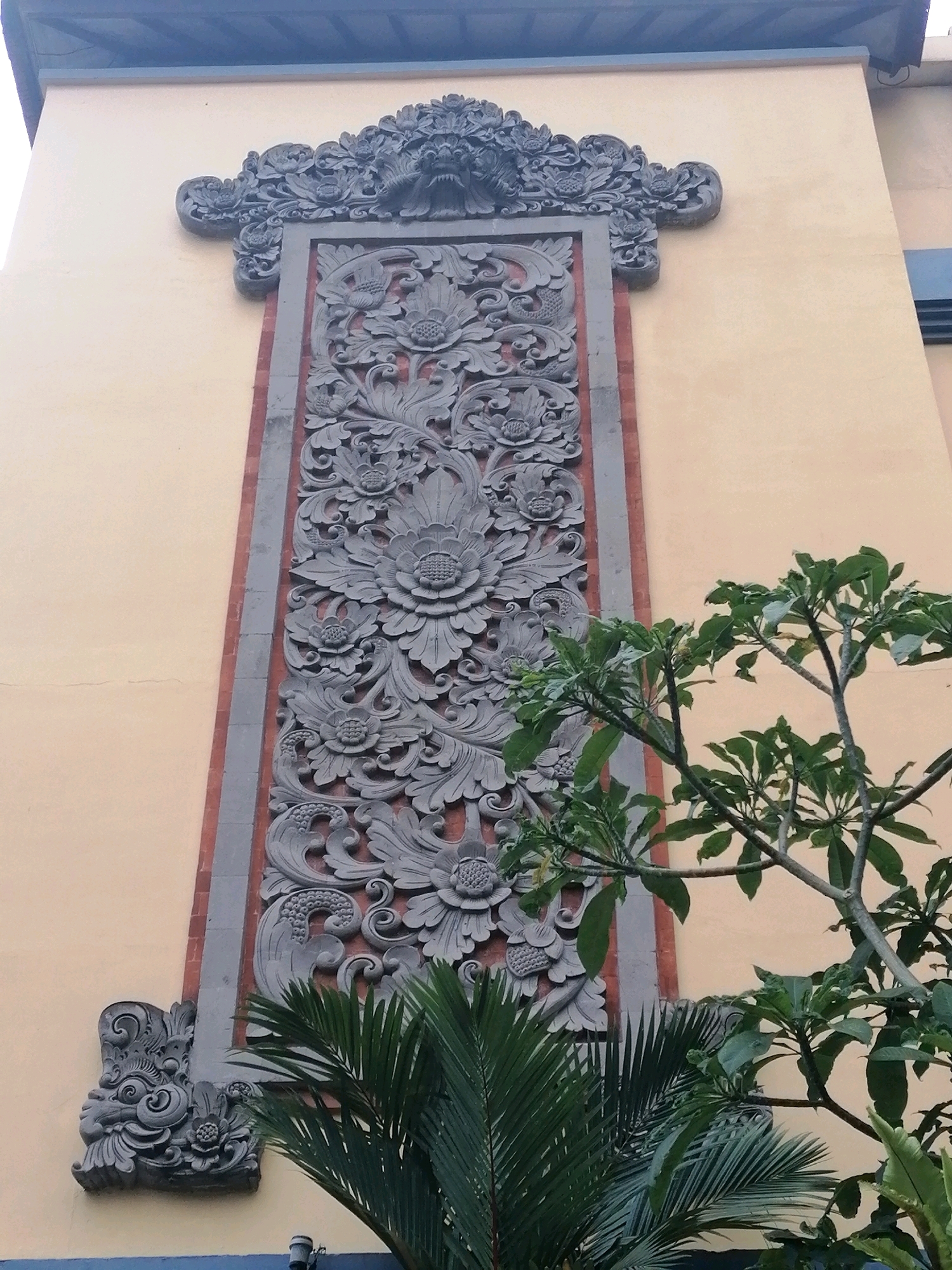 A close-up, low-angle shot captures the ornate architectural details of a building facade in Ubud District, Indonesia. The focal point is a tall, vertical relief carving made of dark stone, intricately decorated with floral motifs and swirling patterns. The carving is set against a textured, pale yellow wall, framed by a reddish-brown brick band and a grey stone border. The top of the carving features a more elaborate, gargoyle-like element with sharp teeth and a fierce expression, flanked by smaller decorative elements.
The lower portion of the image is partially obscured by lush green foliage. A large palm frond, with its characteristic feathery leaves, dominates the foreground. Behind it, branches of a tree with broad, rounded leaves are visible, extending into the frame from the right side. The building's upper edge, with a dark blue trim and a sloped roof, is visible at the very top of the image. The scene is devoid of people. The lighting suggests it is daytime, possibly overcast, as there are no strong shadows. The overall impression is one of detailed craftsmanship and the integration of natural elements with a man-made structure.