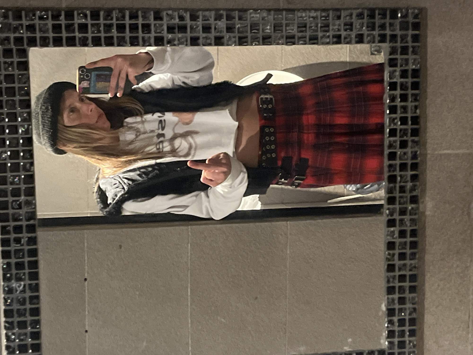 A person is captured in a mirror selfie, reflecting an indoor setting, likely a restroom, given the tiled walls and a partially visible white fixture in the background. The mirror features a distinctive black mosaic tile frame.
The individual, appearing to be a woman with long, light brown hair, is looking directly at the camera while holding up their left hand, extended with the middle finger. She wears a dark grey beanie, a white long-sleeved top with inverted text (appearing as "ЯUGE" or similar), and a black furry or fuzzy vest over it. The lower half of her attire consists of a red plaid skirt adorned with multiple black buckled straps.
She is holding a smartphone, identifiable by its triple-lens camera system on the back, suggesting a recent iPhone model. The phone is encased in a colorful cover with various stickers or designs. The person's facial expression is neutral. The overall lighting suggests artificial indoor illumination. The provided context indicates the location is Buenos Aires, Argentina, though no specific visual cues within the image confirm this.