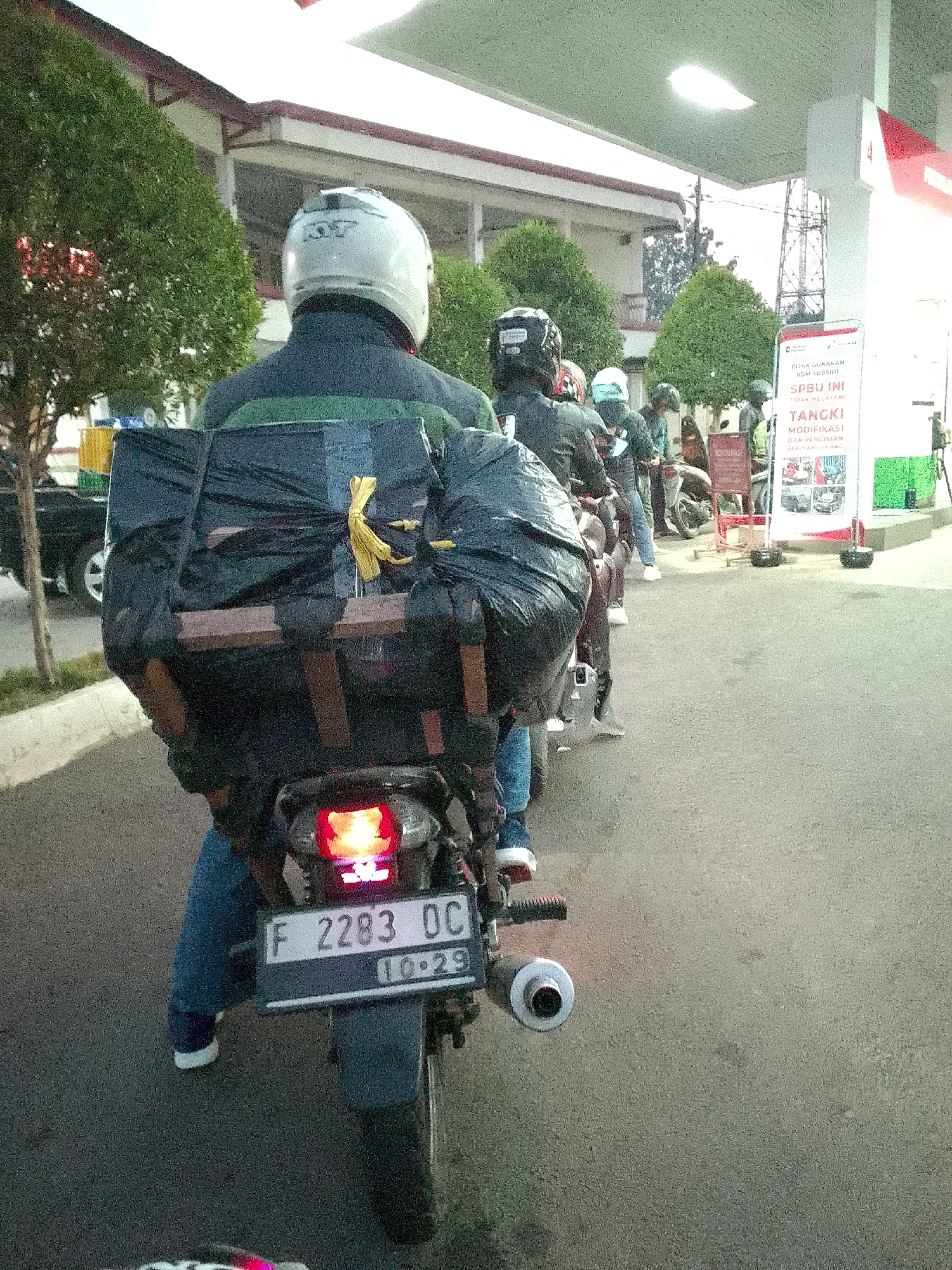 A first-person perspective from a motorcycle shows a large, black, tarp-covered cargo load secured to the rear of the motorcycle. The cargo appears to be a large box with wooden supports and tied down with yellow rope. The motorcycle is in motion, indicated by motion blur. In front, other motorcycles and riders are visible, also appearing to be in motion. A gas station canopy is overhead, and a sign for "SPBU INI TIDAK MELAYANI TANGKI MODIFIKASI DAN PENGISIAN BERUBAH JENIS" (This gas station does not serve modified tanks and changes in fuel type) is on the right. The ground is paved asphalt. The background shows trees, a building, and utility poles. The license plate of the motorcycle reads "F 2283 OC" with "10.29" below it. The scene is likely the gas station in Cipatat, Indonesia.