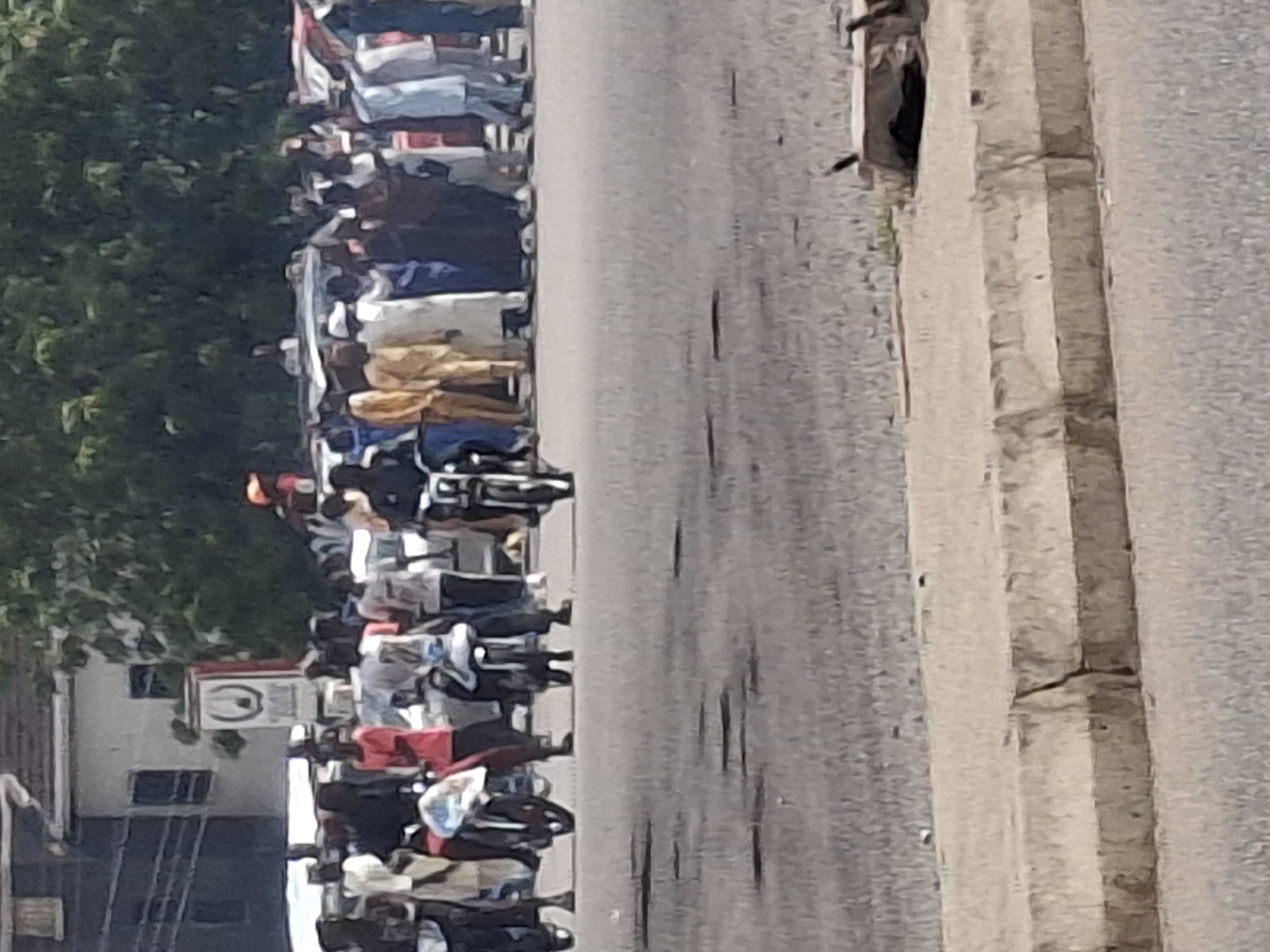 The image depicts a street scene in Gombe, Nigeria, with a crowd of people gathered. Several individuals are on motorcycles, some of which are parked while others appear to be in motion or ready to move. The people are dressed in a variety of clothing, including traditional attire and casual wear. In the background, there is a building with a sign and trees. The ground appears to be paved, with a concrete barrier in the foreground. The overall impression is of a busy public area with ongoing activity.