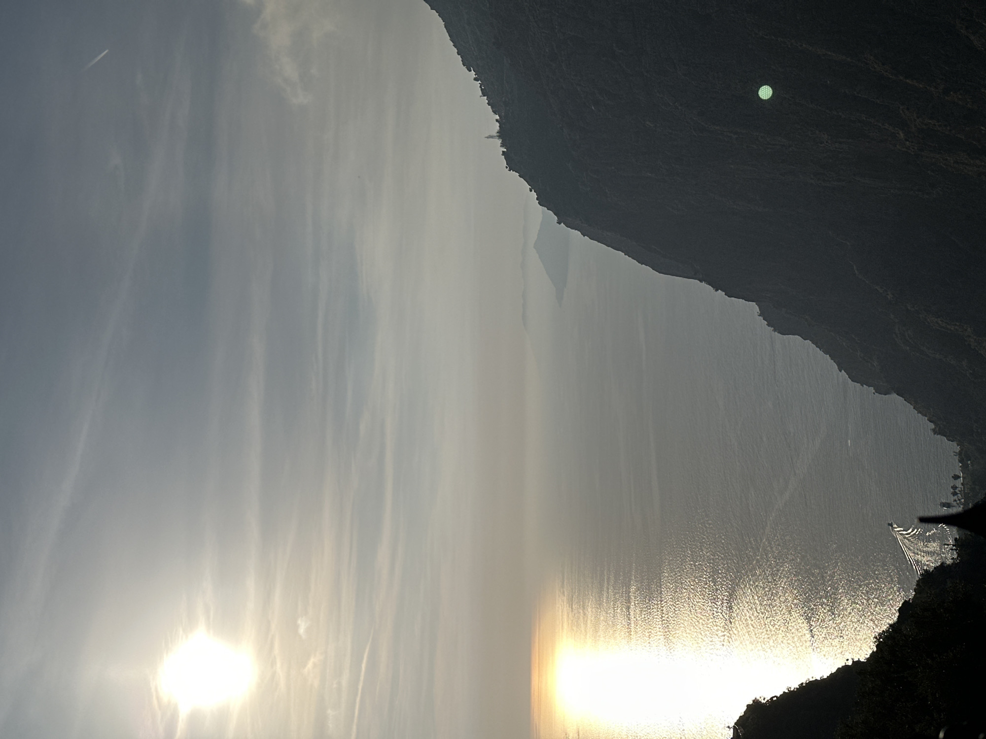 The image captures a serene, expansive landscape, likely during sunrise or sunset, overlooking a large body of water framed by towering cliffs and mountains. The scene is oriented vertically, showing the sky at the top, the sea in the middle, and landmasses on the right and bottom.
The sky, occupying the upper left portion, is a soft, hazy blue-gray, streaked with wispy clouds. A bright, diffused sun or strong lens flare is visible in the upper left, casting warm light across the sky. A faint white contrail from an aircraft is discernible against the sky.
The central part of the image is dominated by a vast body of water, shimmering with intense reflections of the low-lying sun. The surface of the water shows numerous ripples and patterns of light, creating a glistening effect. Along the bottom right edge of the water, a wake, possibly from a moving boat, creates V-shaped patterns leading away from a dark, indistinct object or structure near the shore.
Dark, rugged cliffs and mountains form a dramatic silhouette along the right side and bottom of the frame. Their steep, textured surfaces appear in shadow. High on a distant cliff line, a small, distinct white building or structure is visible. Against the dark, textured surface of the foreground cliff, a prominent, luminous green circular light or reflection stands out.
Given the location context of Fethiye, Turkey, this is likely a view of the Mediterranean Sea or a large bay surrounded by the region's characteristic mountainous coastline. The low angle of the sun and the warm, diffused light indicate either early morning or late afternoon. No discernible text is visible in the image.