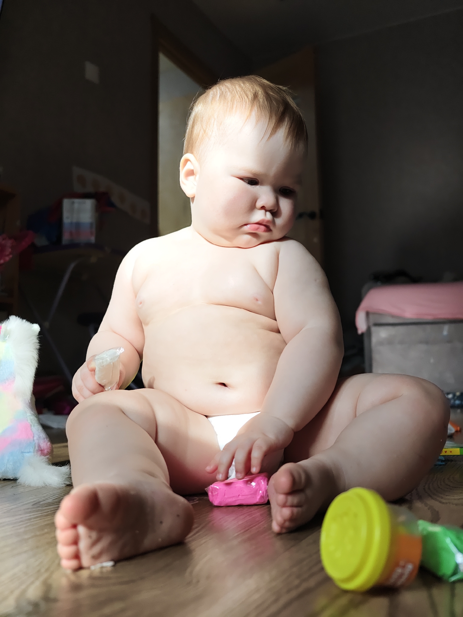 An infant, approximately 9-18 months old with light hair and fair skin, is seated cross-legged on a light brown wooden floor. The infant is shirtless, wearing a white diaper, and looks downward towards the right. The left hand holds a small, clear plastic object. The right hand rests near a pink, rectangular toy. To the infant's immediate left, a portion of a white, fluffy toy with pastel rainbow accents is visible. On the floor to the right are a yellow cylindrical container with an orange label and a green object. The background is dimly lit, featuring a dark wall, a dark wooden door frame, and indistinct furniture including a bed with a pink blanket on the right, and shelving on the left where a white box with red and blue stripes is visible. The scene is illuminated by strong overhead lighting, creating shadows in the background. This indoor residential scene is situated in the city of Chaikovsky, Russia.