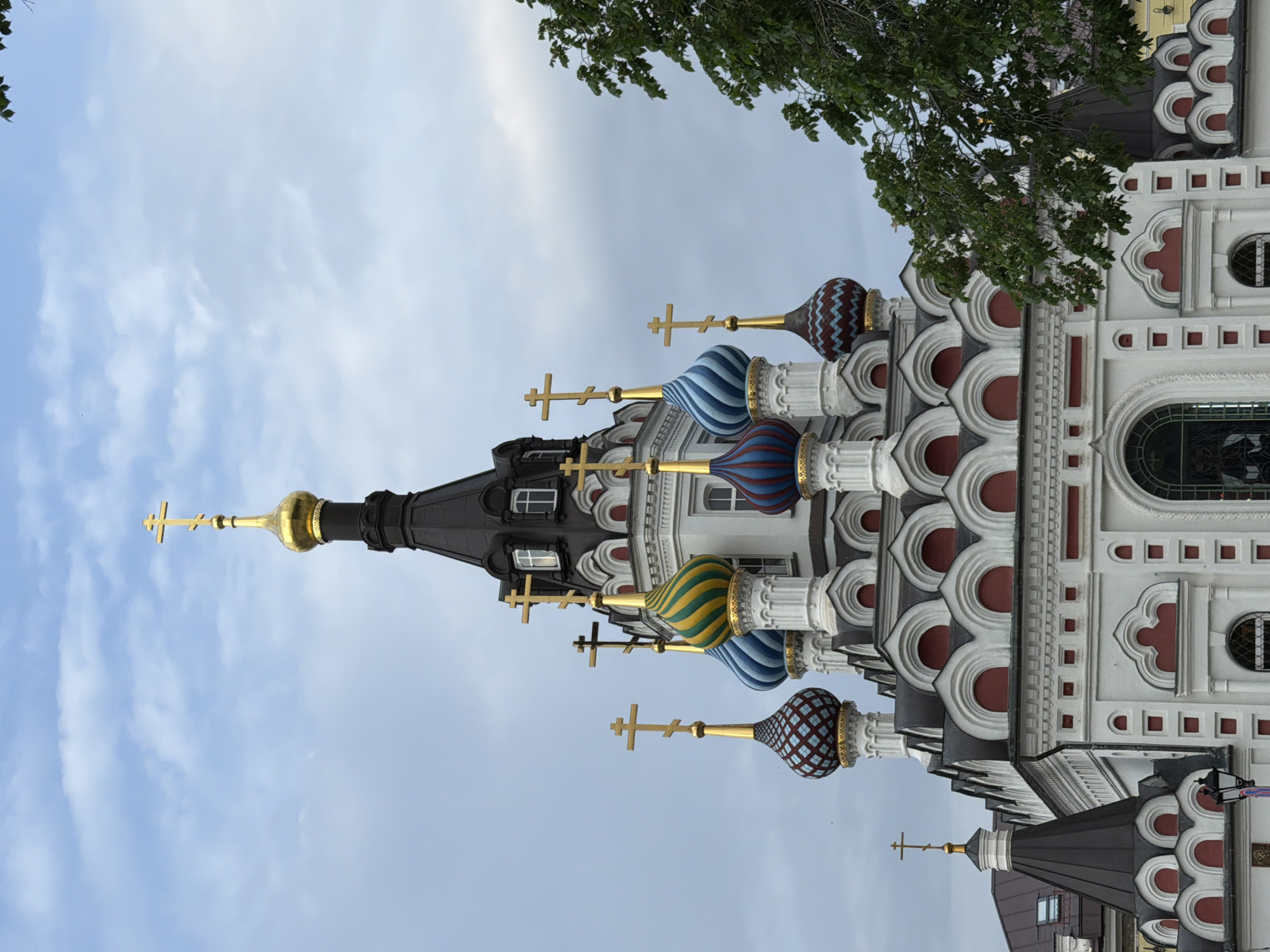 The image shows the Holy Trinity Cathedral in Saratov, Russia, from a low angle. The building is oriented vertically with the domes pointing upwards. The architecture is elaborate, featuring multiple colorful onion domes adorned with golden crosses. The building's facade is primarily white with red and gray accents. The sky is visible in the background, with light clouds suggesting a cloudy day. Green foliage is present at the top right corner of the frame, suggesting a tree. The overall scene captures the architectural detail and religious significance of the cathedral.